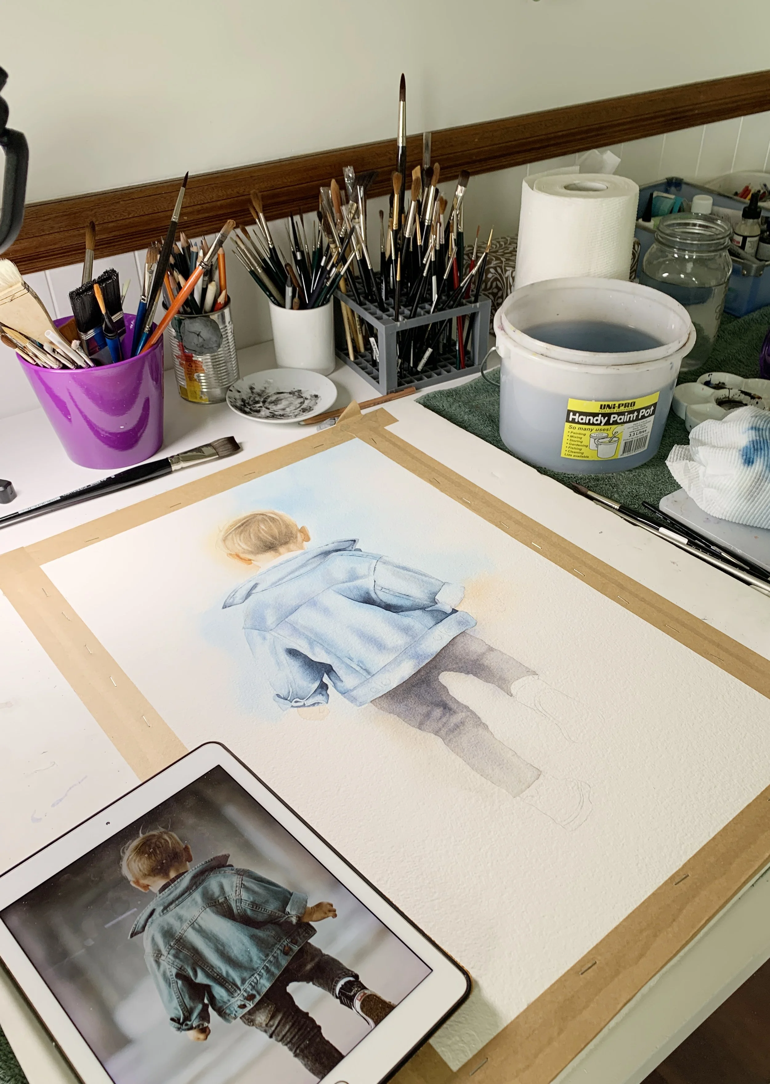 A watercolour painting in progress showing a young boy in a blue denim jacket walking away, with the reference photo on an iPad.