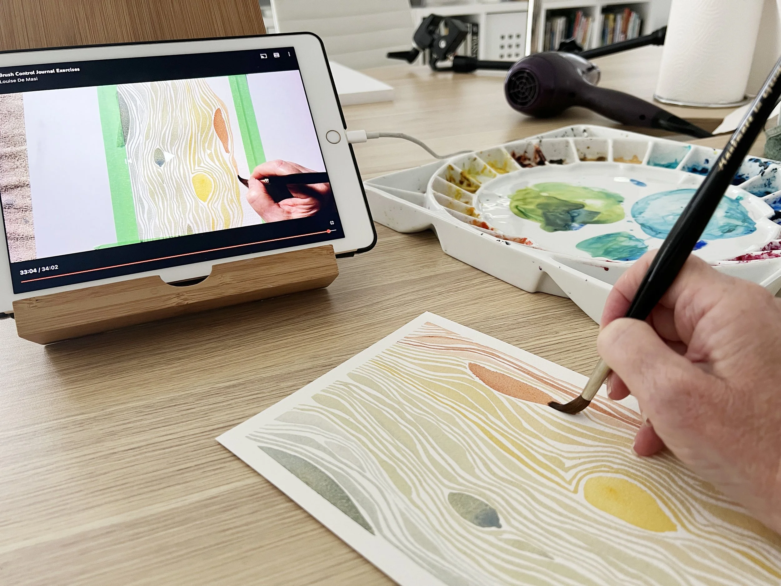 Following a watercolour brush control tutorial with a reference video playing on an iPad and painting along with abstract woodgrain-style lines.