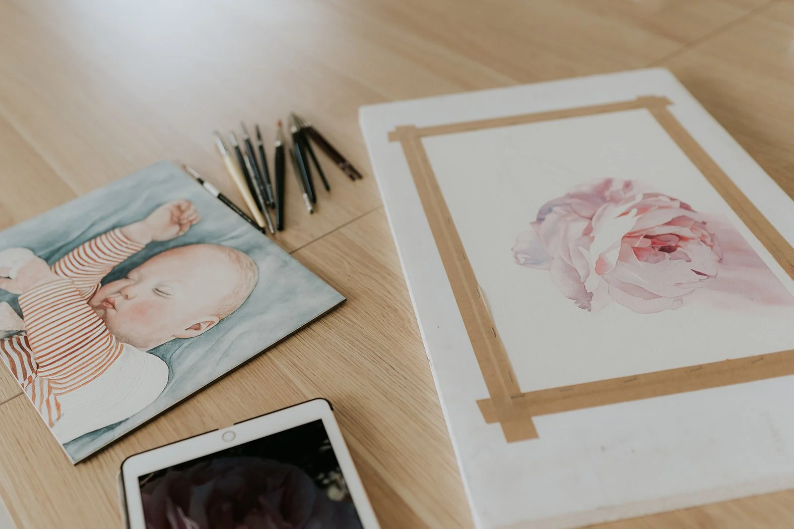 A watercolour painting of a sleeping baby and a partially finished rose lay on a wooden surface surrounded by brushes and an iPad reference image.