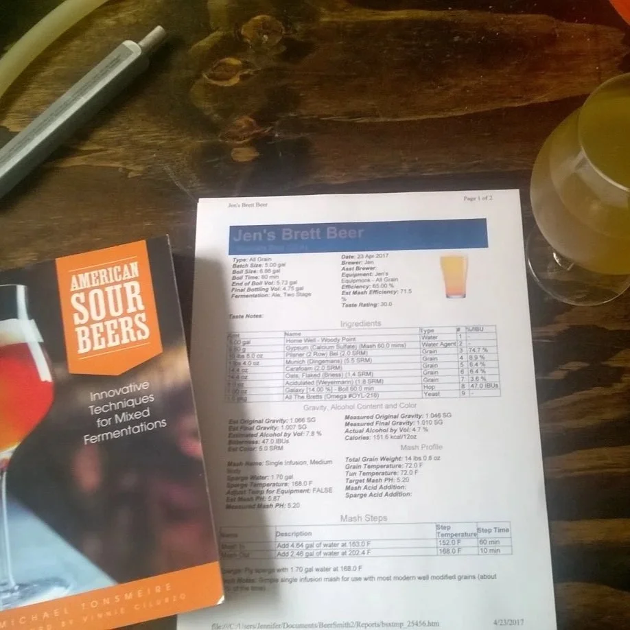 Beer recipe named Jen's Brett Beer with American Sour Beer book and glass of light-colored beer on wooden background