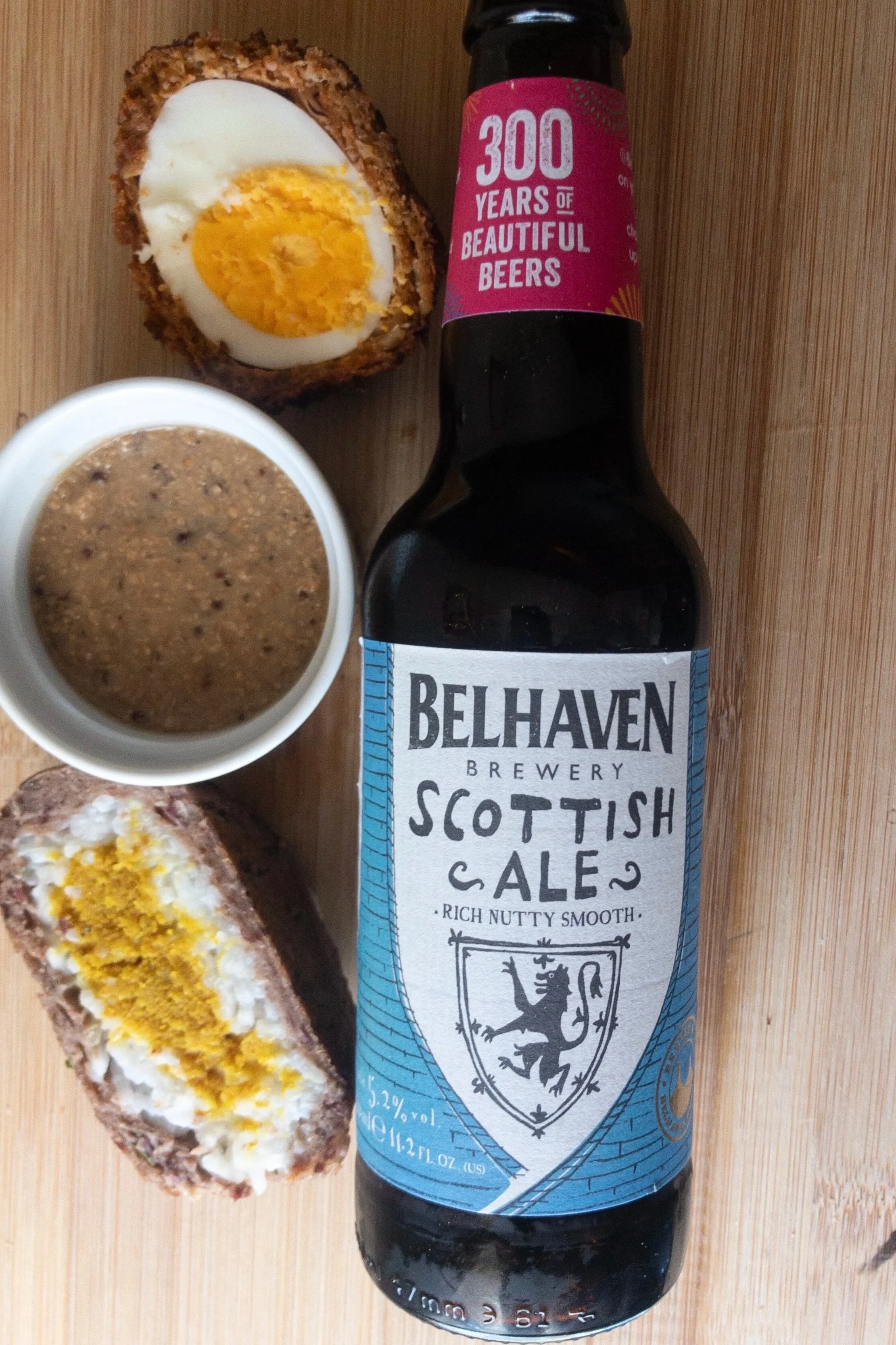 Scotch eggs cut in half with white container of mustard next to Belhaven Scottish Ale bottle on wood background