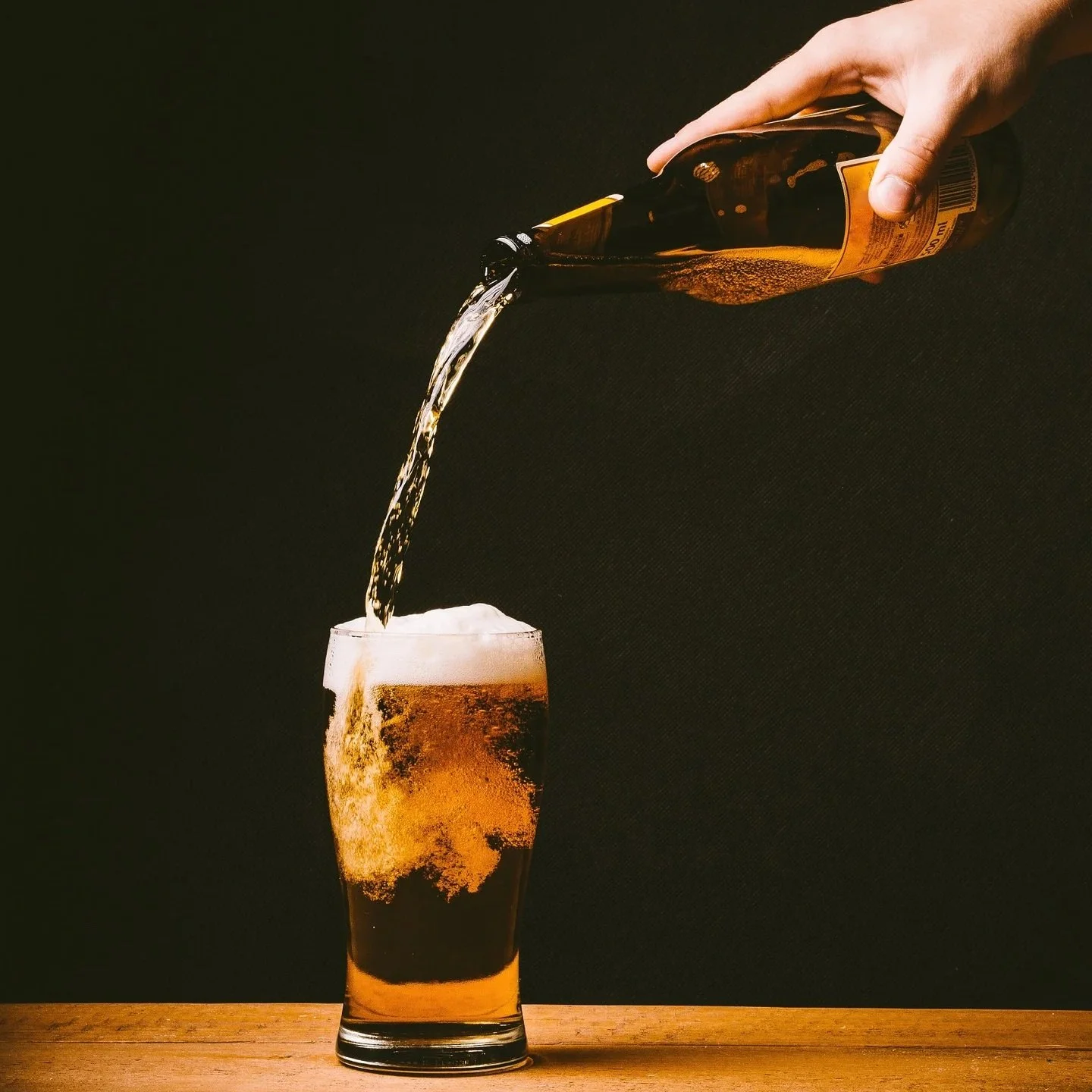 White hand holding brown beer bottle pouring beer into glass sitting on a wood table