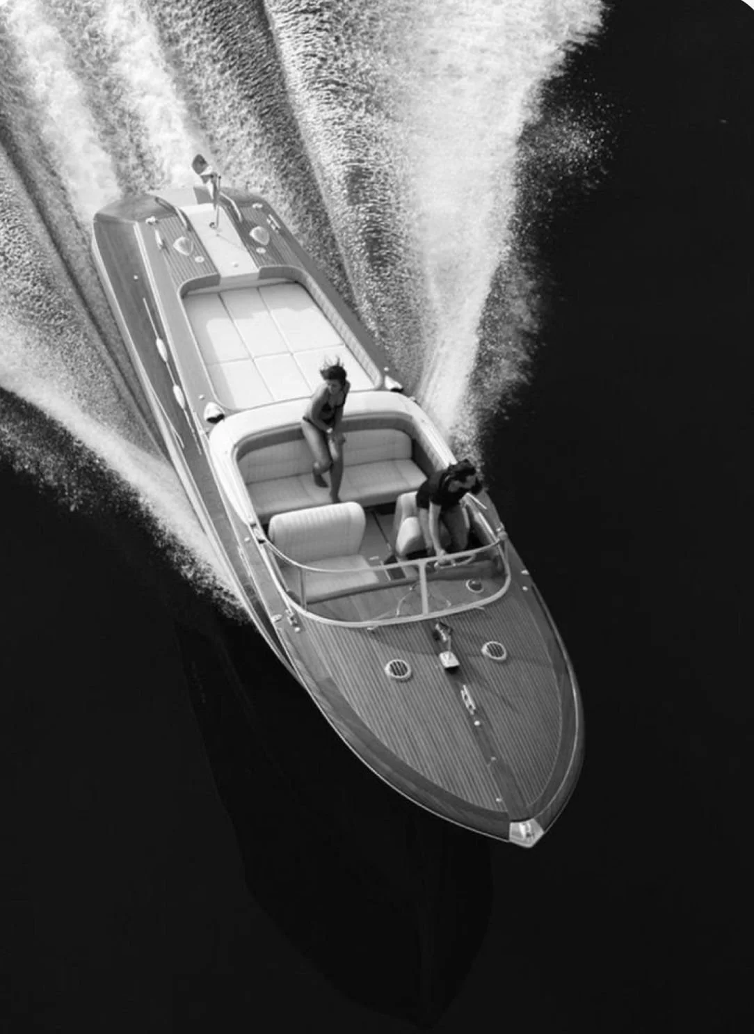 A black-and-white aerial photo of a yacht sailing in water, with two women on deck, one standing and the other sitting near the helm.