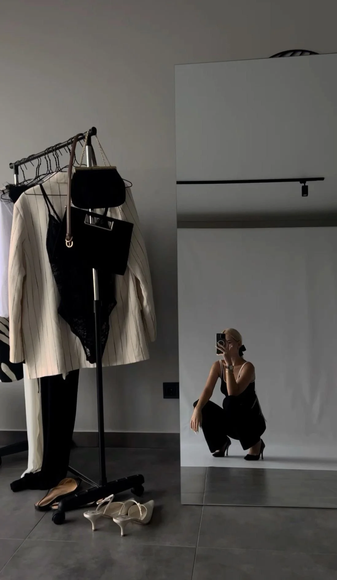 A woman taking a mirror selfie, squatting and wearing black high heels, a black top, and black pants, with a white background and clothing on a rack beside her.