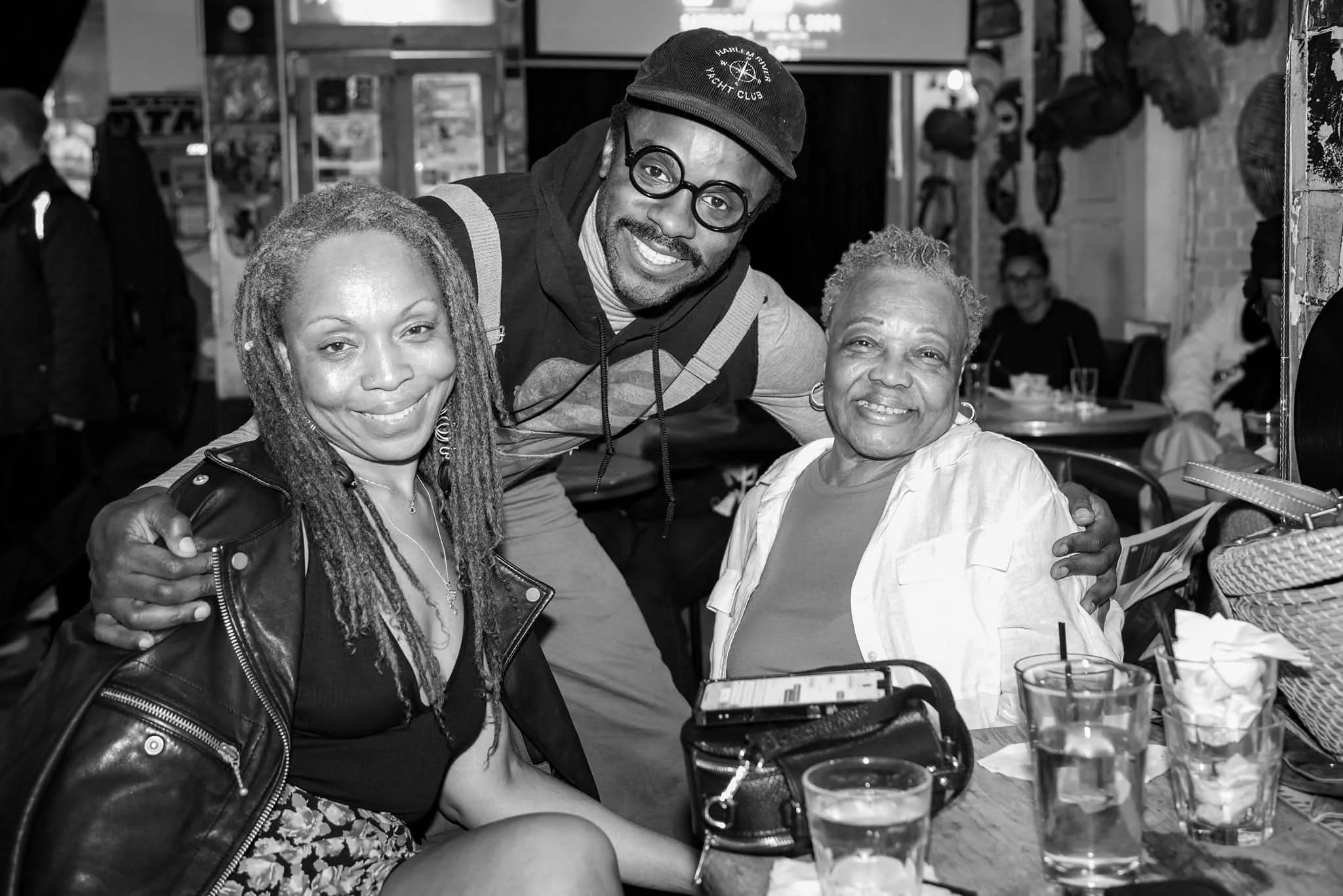 Three people sitting at a table in a restaurant, smiling at the camera. A man with glasses and a hat is standing behind two women, one with dreadlocks and the other with short, curly hair. Drinks are on the table.