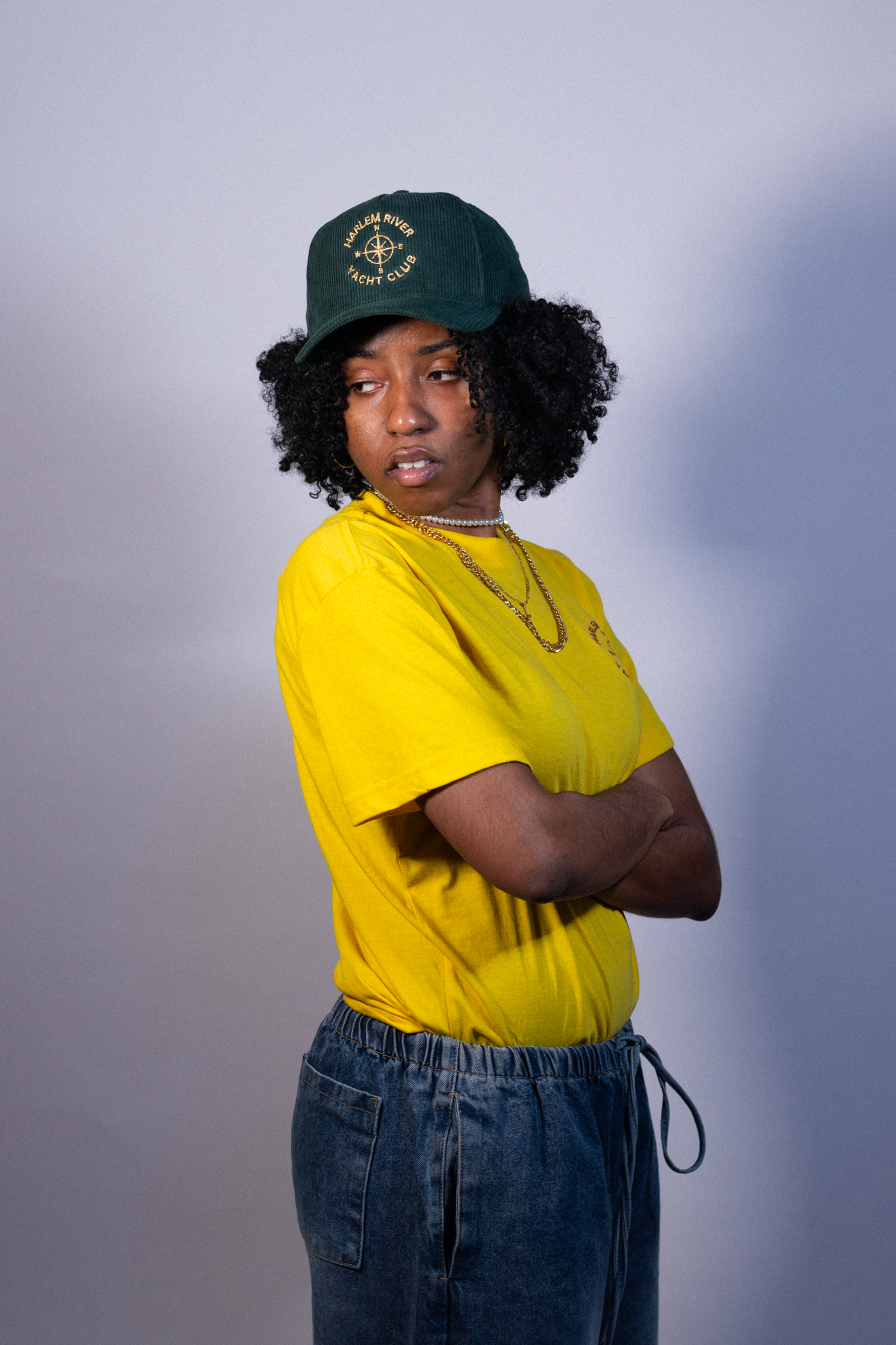A young woman with curly black hair wearing a yellow t-shirt, a green cap with yellow embroidery that says 'Harlem River Yacht Club,' layered necklaces, and dark jeans, standing against a plain light gray background, with her arms crossed and looking