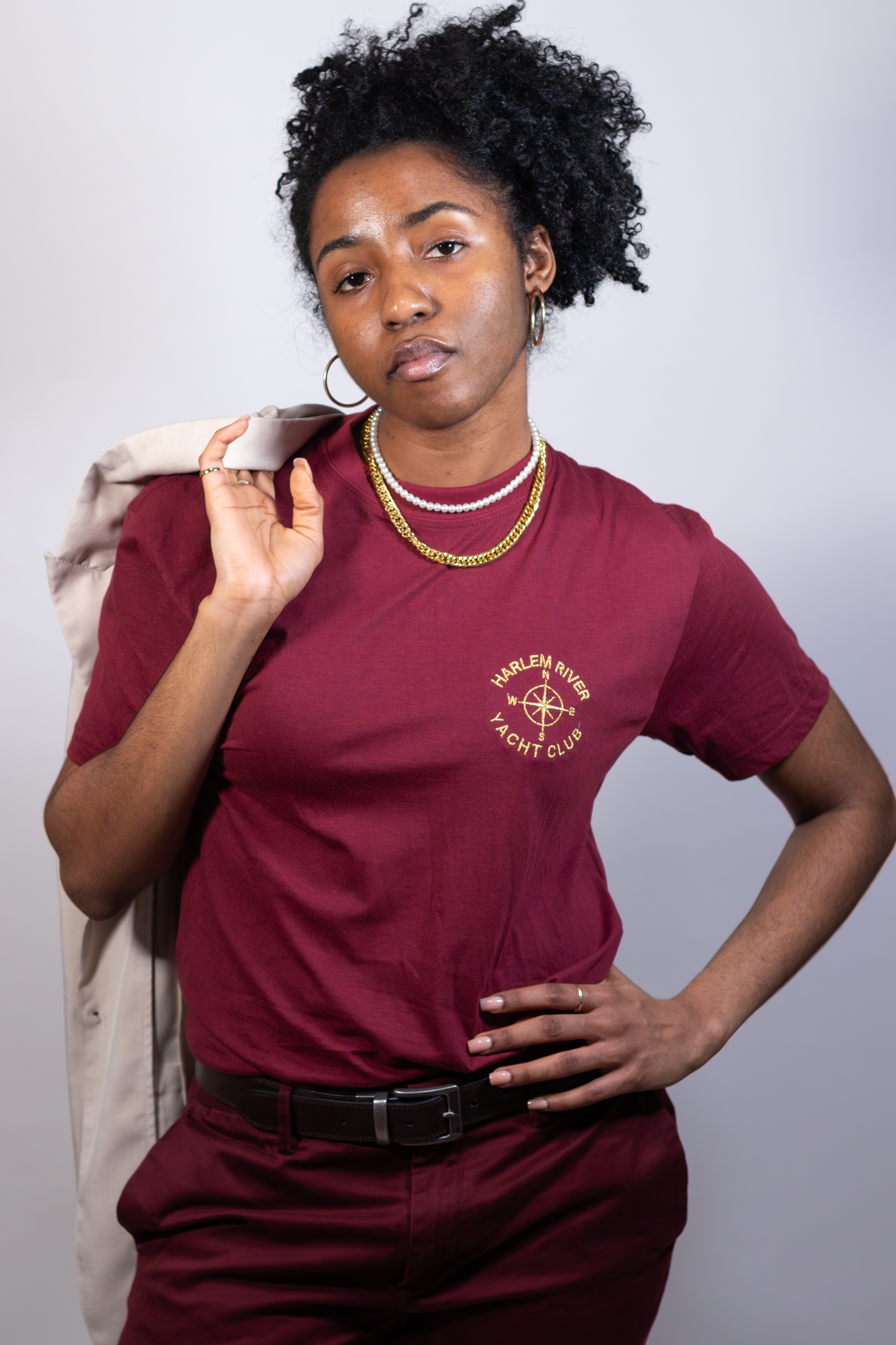 A woman with short curly black hair, wearing a burgundy t-shirt with 'Harlem River Yacht Club' and a compass logo, accessorized with gold and pearl necklaces, hoop earrings, and a ring, stands against a plain white background, holding a light-colored