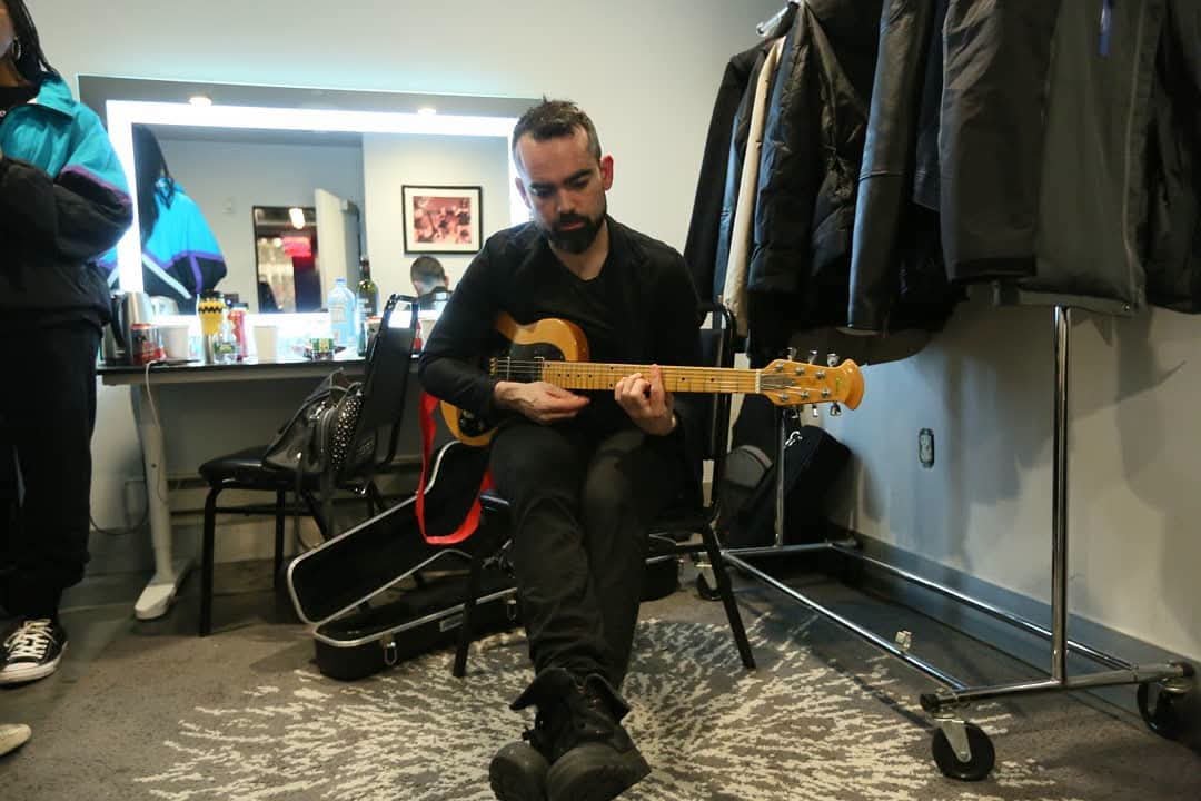 A man sitting on a chair, playing an electric guitar in a room with jackets hanging on a rack, a table with snacks and drinks, and a large mirror behind him.
