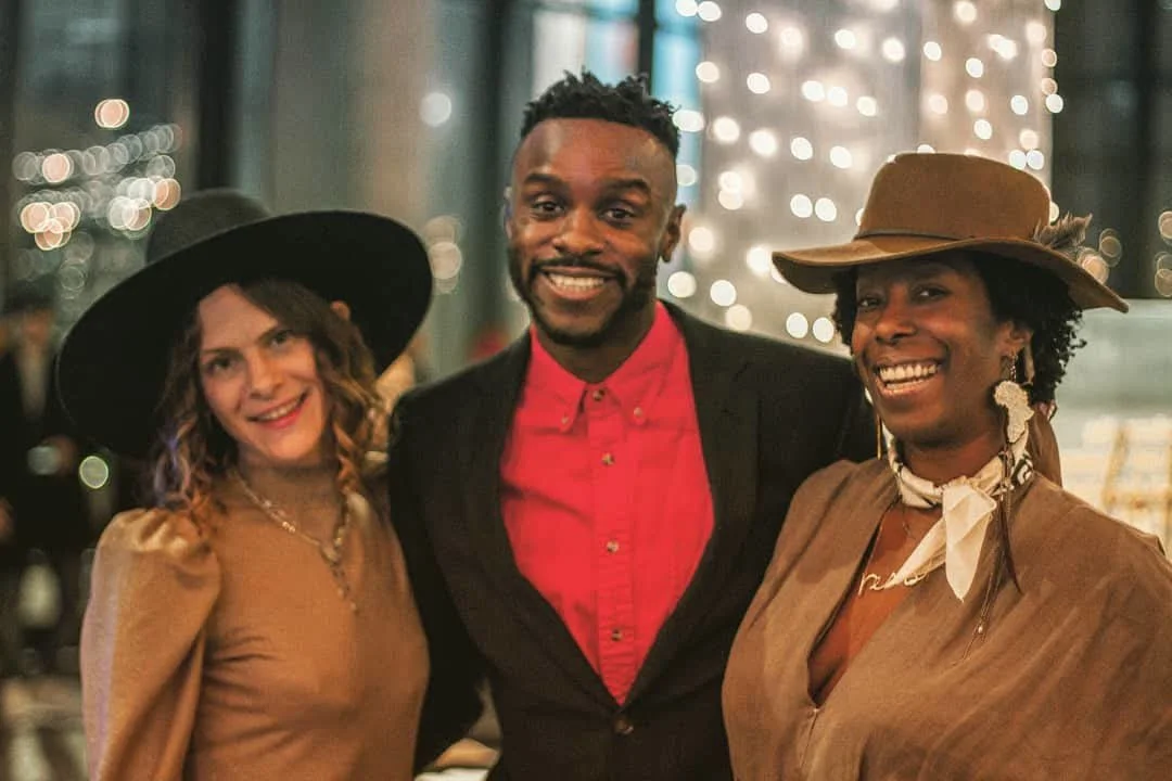 Three people smiling inside a restaurant with blurred lights in the background. Two women on either side of a man, all dressed in stylish, vintage-inspired clothing with hats.