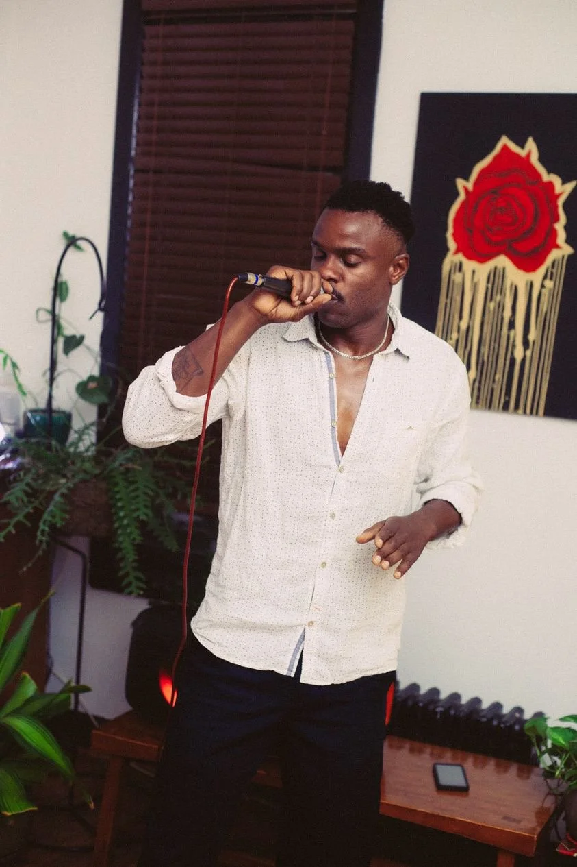 A man with short black hair singing or speaking into a microphone indoors, wearing a white button-up shirt with the top buttons undone and black pants, with plants and artwork featuring a red rose and dripping gold paint in the background.