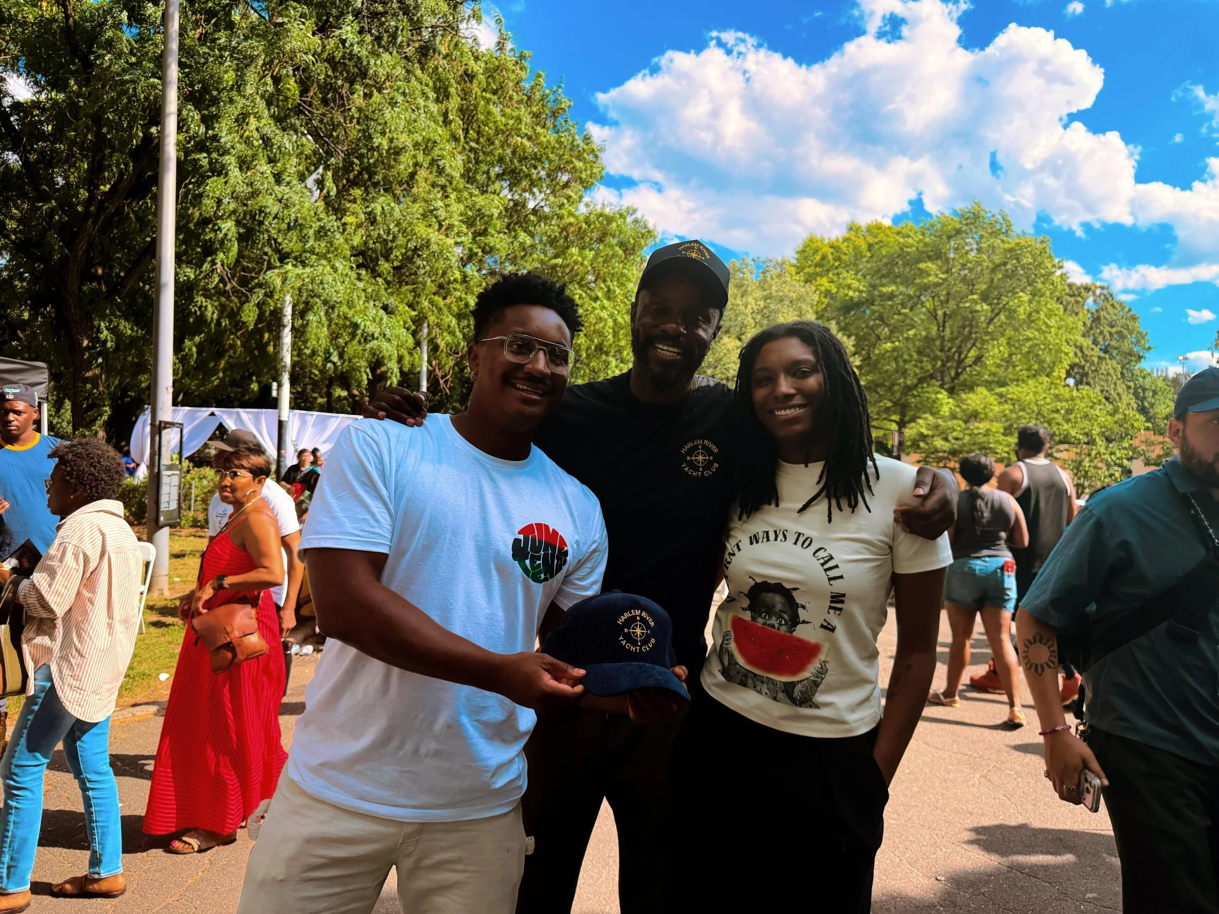 Three people standing together outdoors at a park or festival, smiling at the camera. There are trees, a blue sky with clouds, and other people in the background.