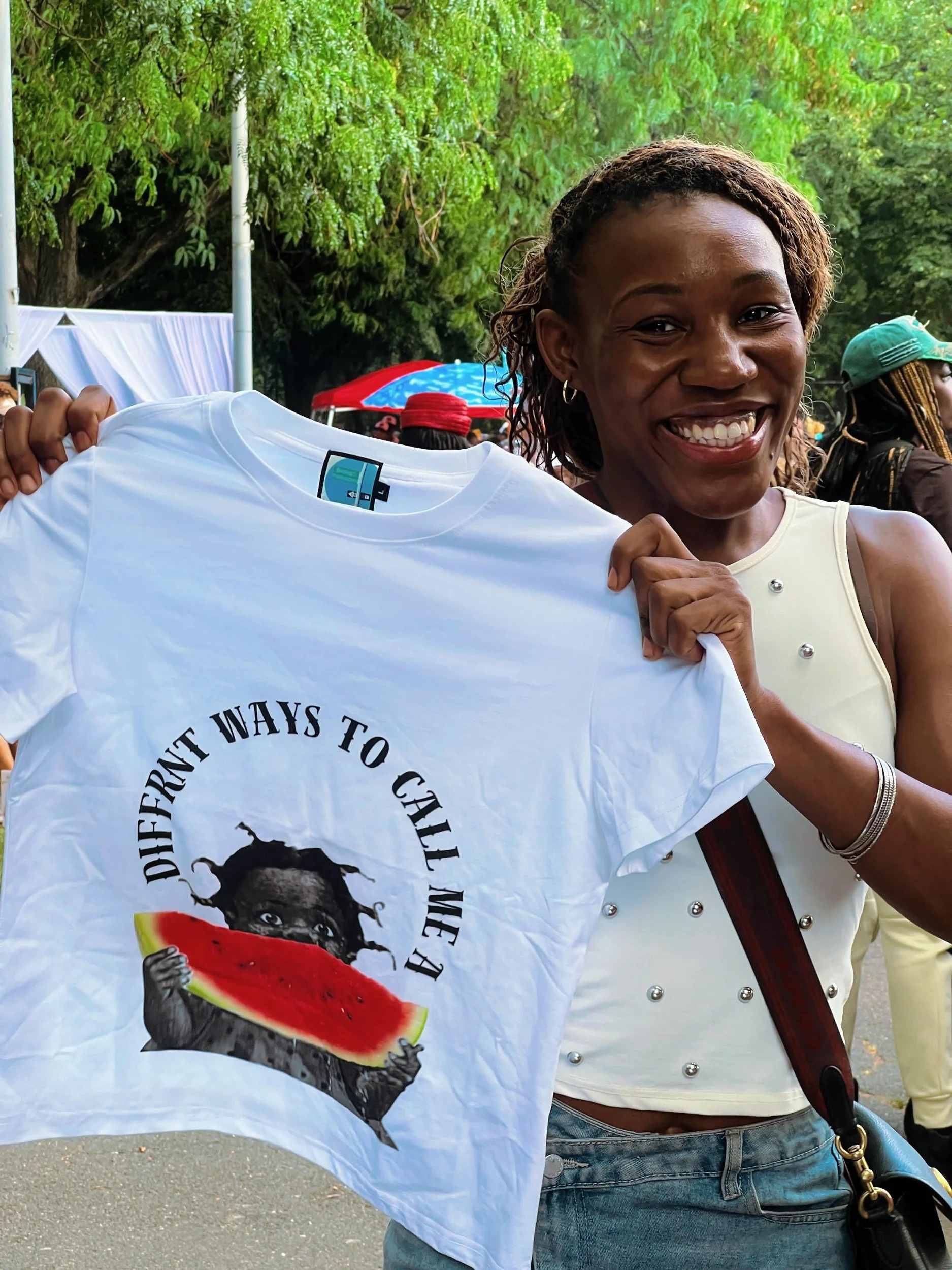 A smiling woman with braided hair and hoop earrings holds a white t-shirt with a graphic design of a woman holding a slice of watermelon, with the text 'Different Ways to Call Me' on it. She is outdoors, with green trees and other people in the backg