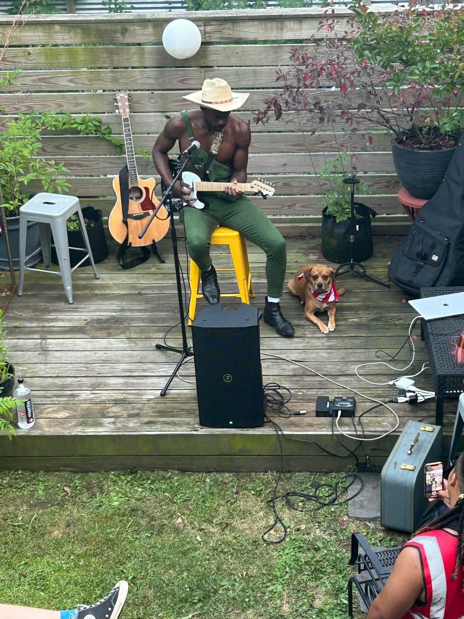 A shirtless man wearing a wide-brimmed hat playing an electric guitar and sitting on a yellow stool. There is a dog lying on the grass next to him. The background shows a wooden fence with plants and a large potted plant on a stand.