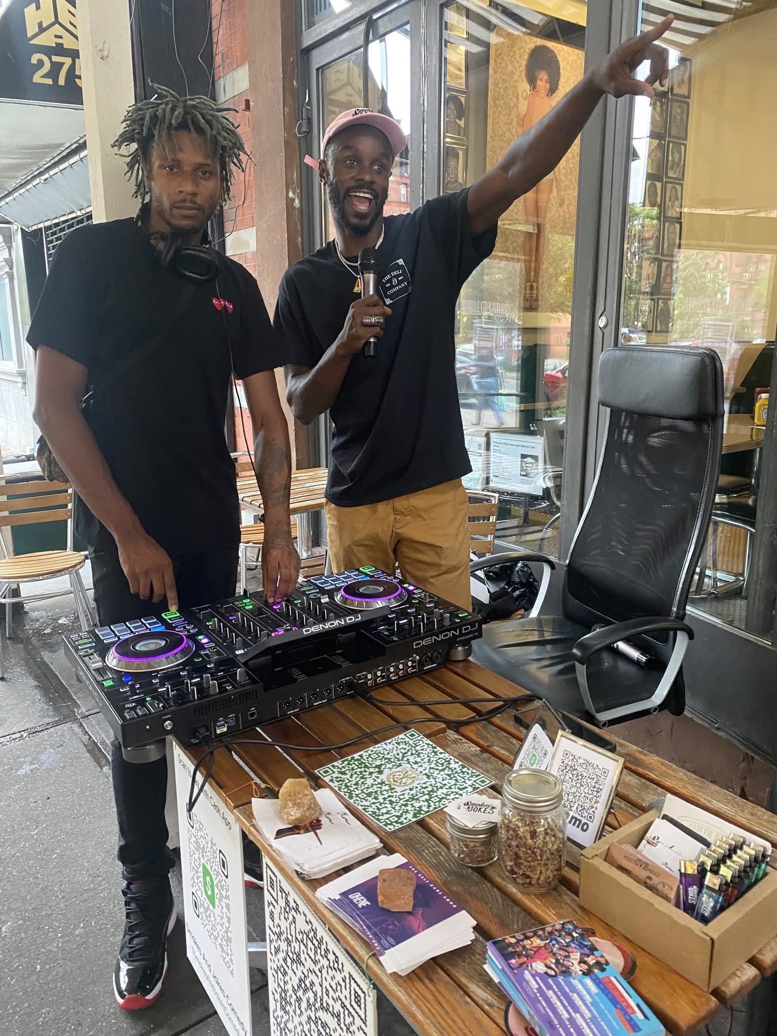 Two men are standing behind a table with a DJ controller, flyers, and QR codes. One man is holding a microphone and pointing upwards. There are a chair, jars, and promotional materials on the table. The background shows a glass window and some artwor