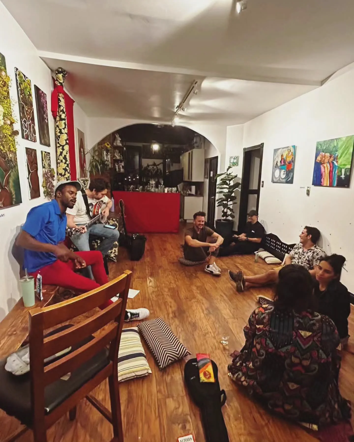 A group of people, some sitting and others standing, enjoying a musical gathering in a cozy room with white walls decorated with paintings and hanging plants. One person is playing a guitar, and others are engaged in conversation or listening. The ro