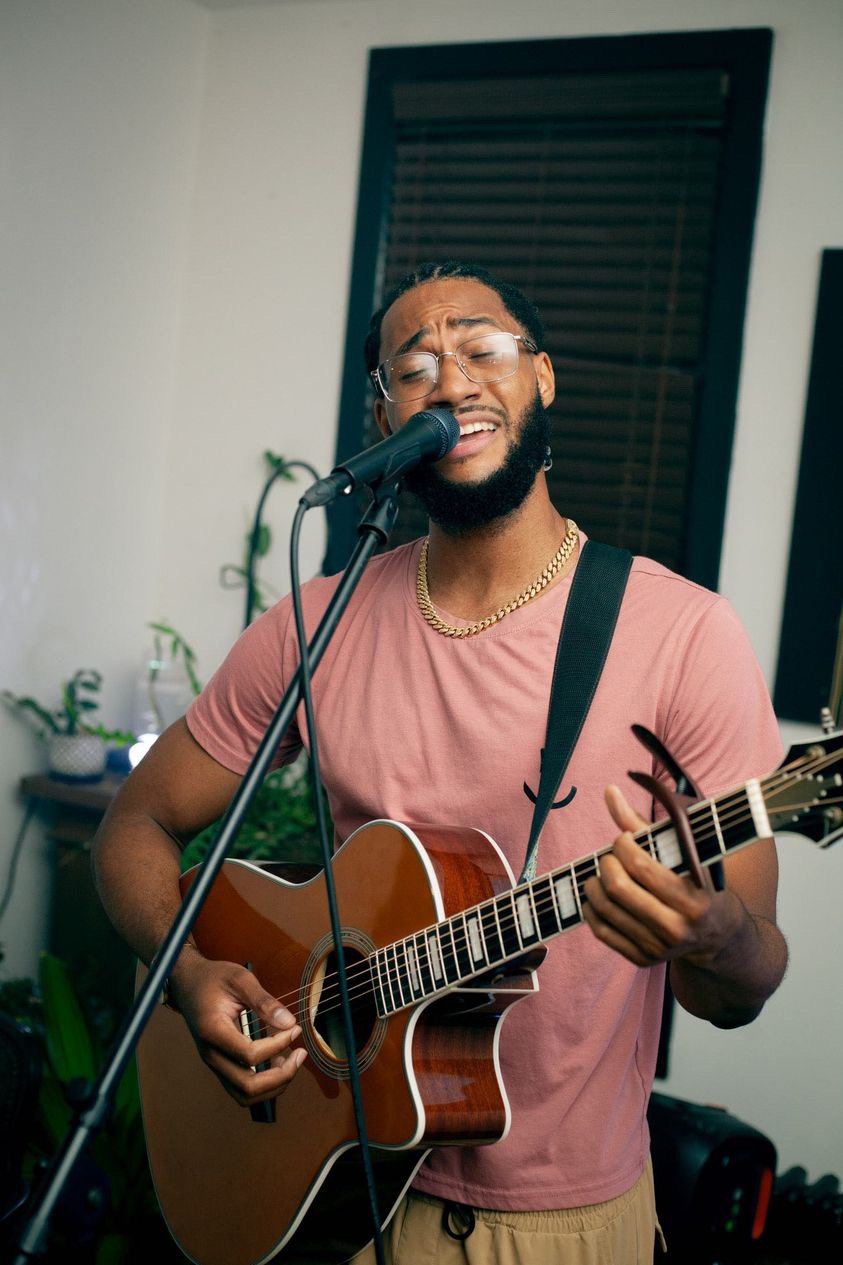 A man with glasses, a gold chain, and a beard, singing passionately into a microphone while playing an acoustic guitar in a room with a window and plants.