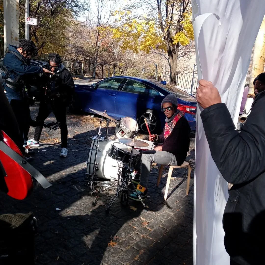 Man playing drums outdoors, surrounded by a camera crew and onlookers, with a blue car and autumn trees in the background.
