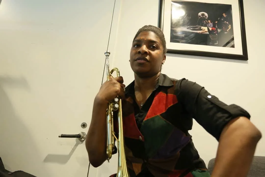 A woman holding a trumpet, sitting in a room with a framed picture on the wall behind her.