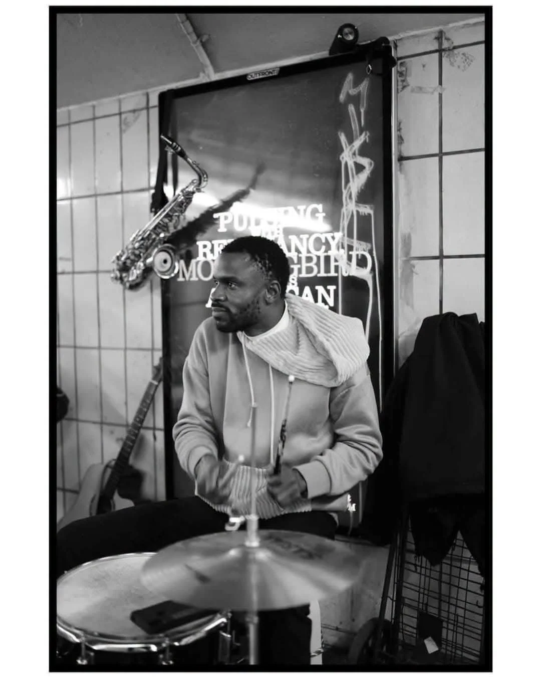 A Black man sitting behind a drum set with a saxophone hanging on a stand beside him, in a room with tiled walls and a neon sign in the background. The image is in black and white.