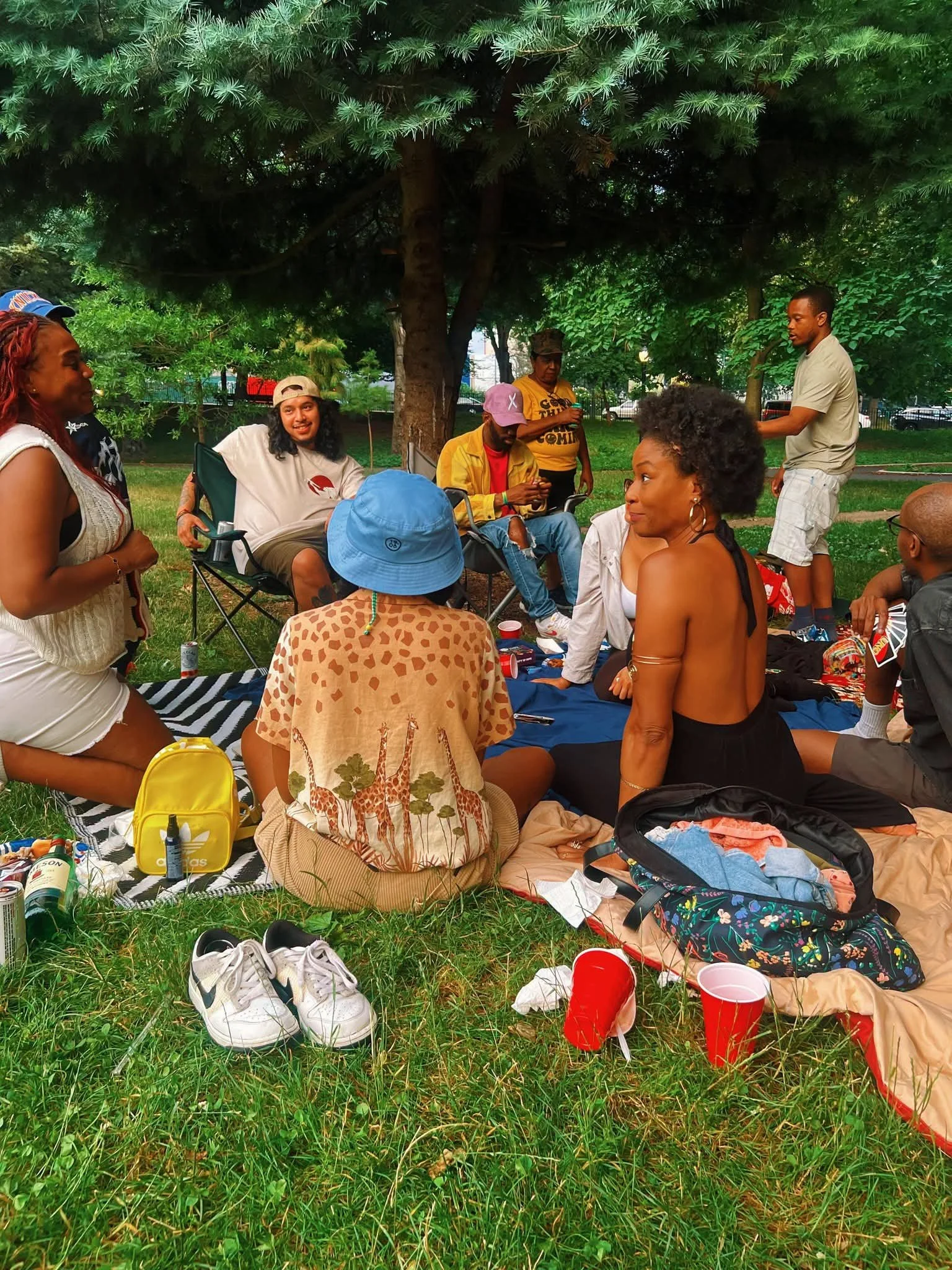 Group of diverse people having a picnic outdoors under a large tree with grass and picnic supplies on the ground.