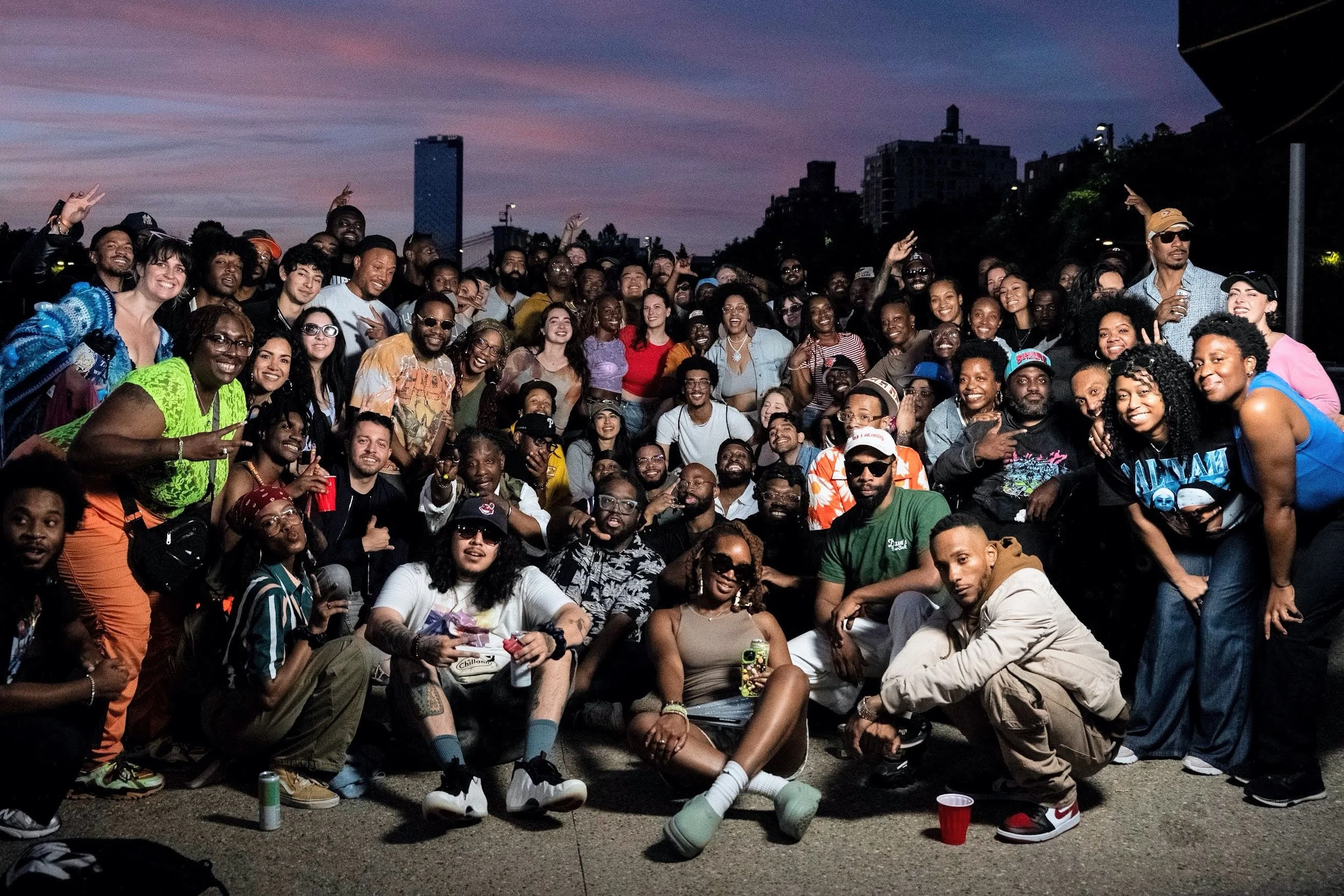 A large group of people gathered outdoors during dusk, smiling and posing for a photo, with city buildings and a bridge in the background.