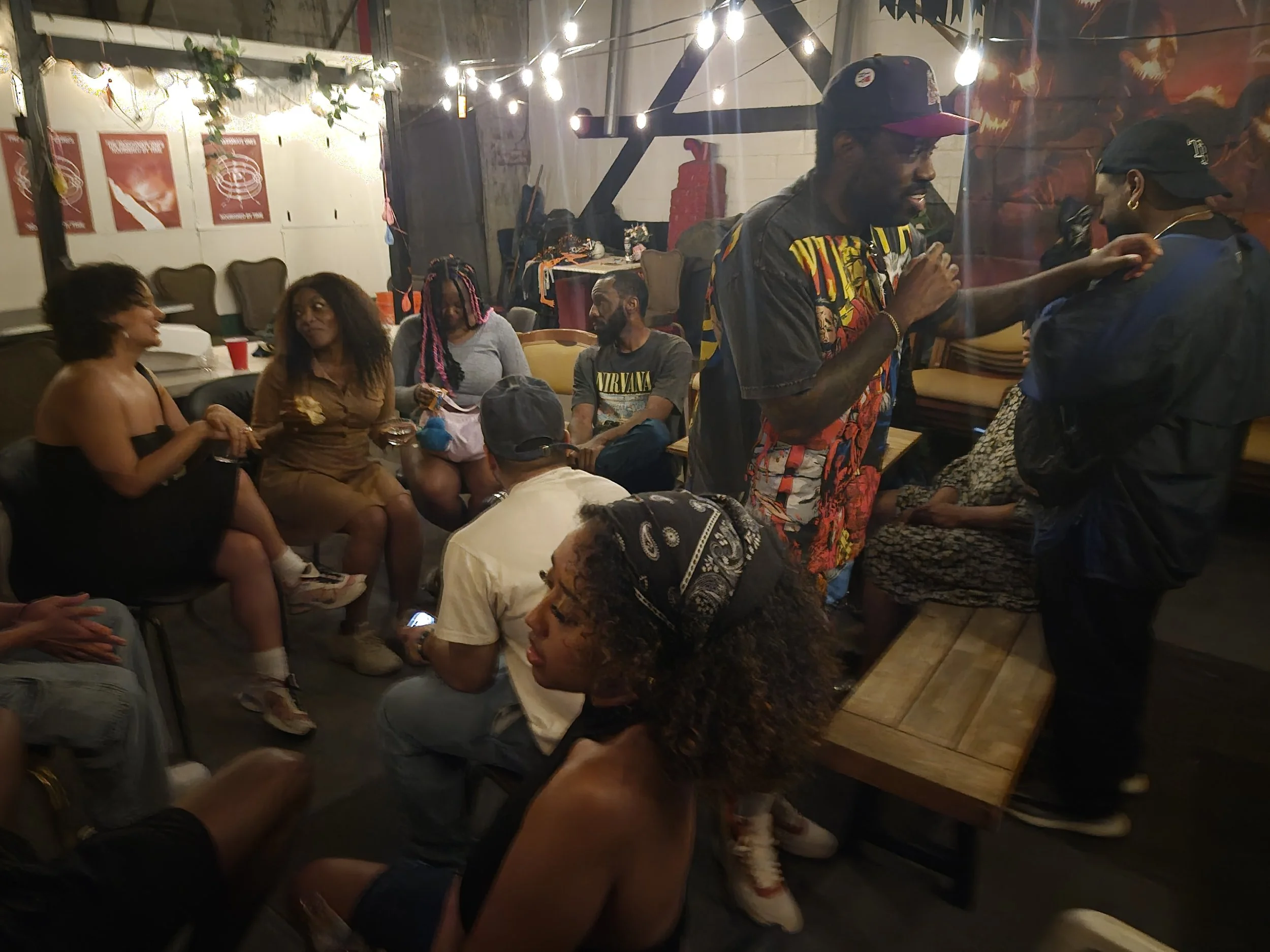 Group of friends socializing in a dimly lit room; some are sitting while others are standing, engaging in conversation. The room is decorated with string lights and posters on the wall, creating a cozy atmosphere.