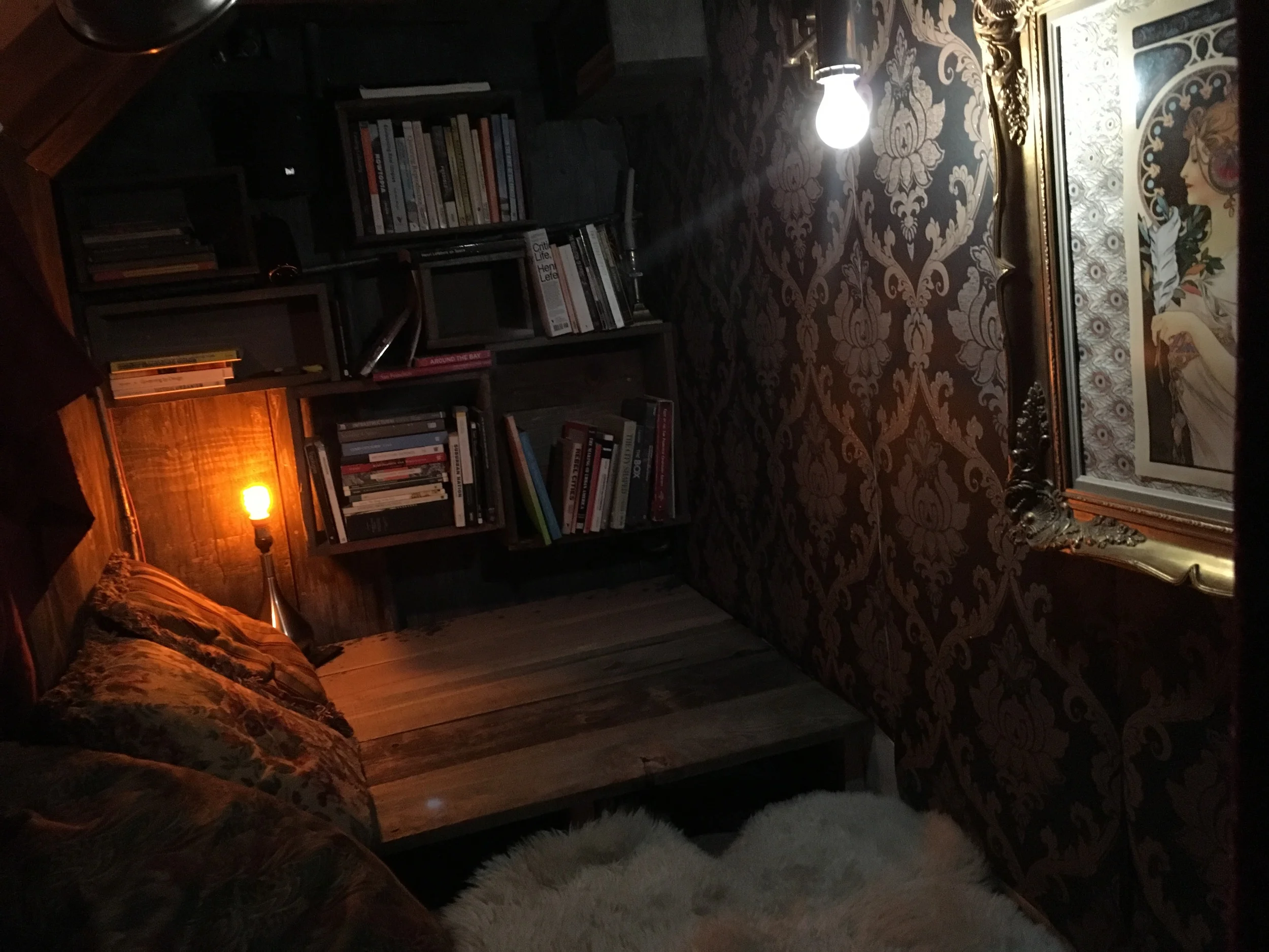 after - recycled redwood fenceboards make up the floor and shelves, with vinyl damask wallpaper, Goodwill-sourced throw pillows, a shag rug, an old picture frame with an Art Nouveau poster, and dim lighting. 