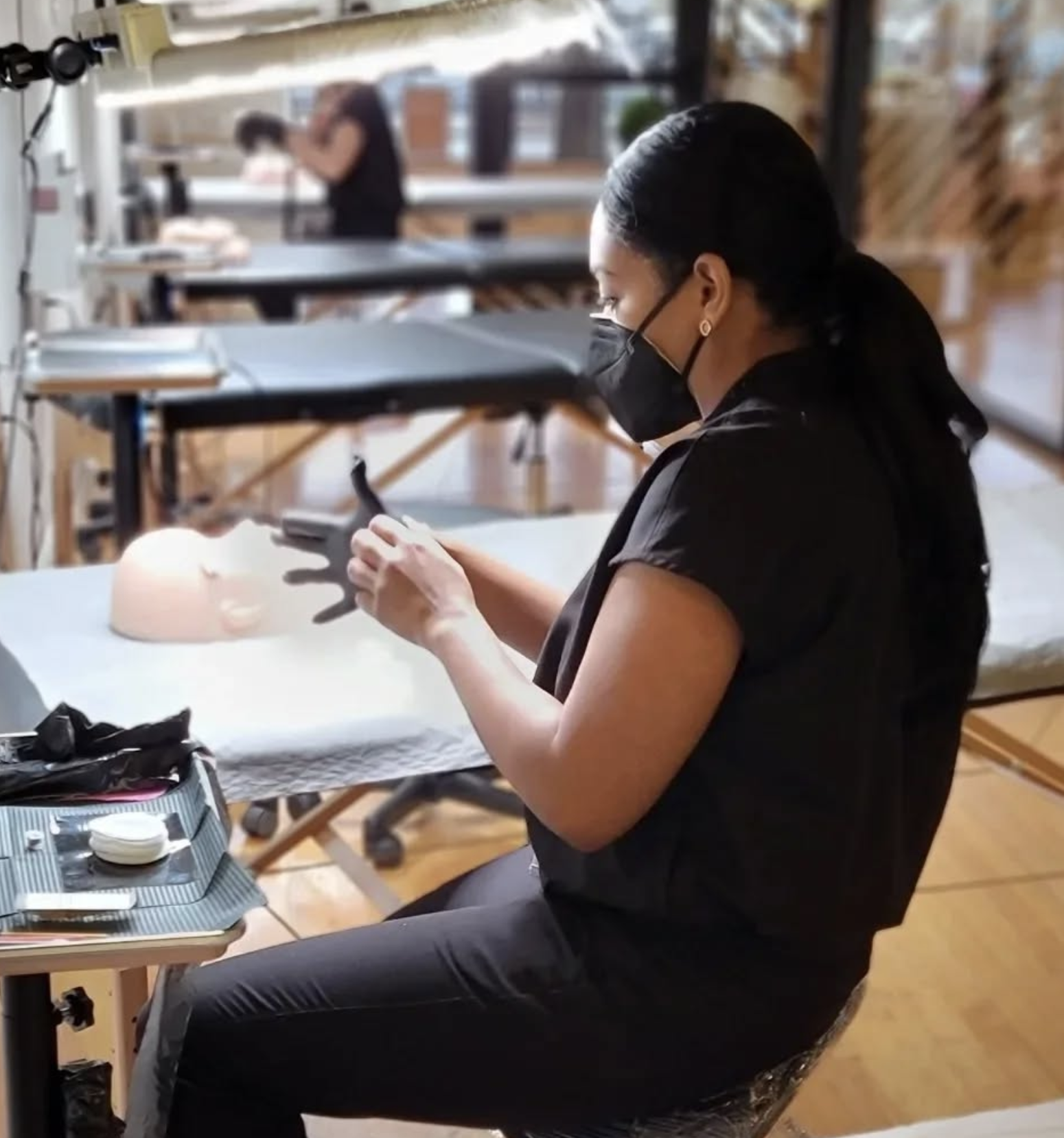 Woman wearing a black mask and black scrubs, seated at a table. She is in a workspace with multiple tables and another person working in the background.