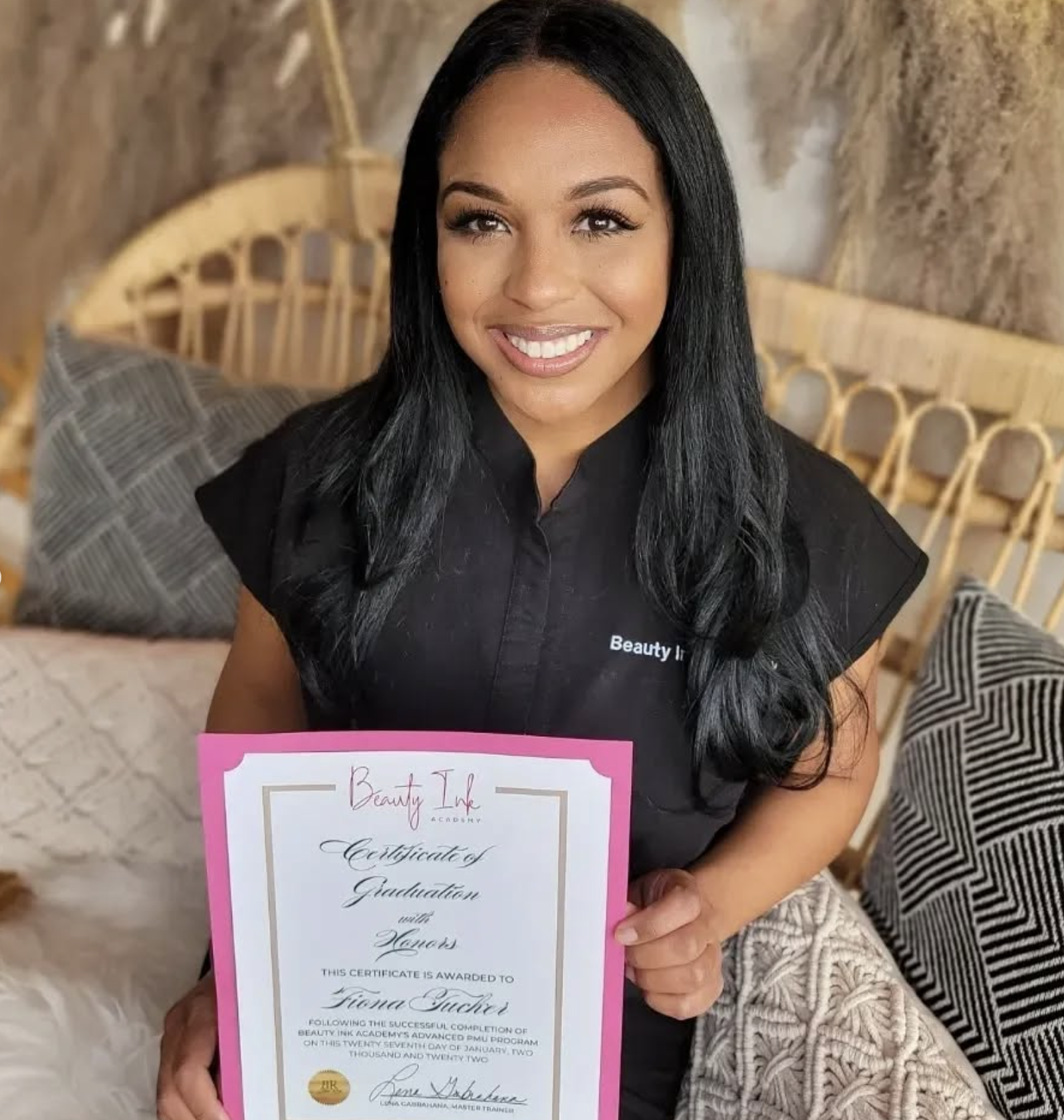A woman with long black hair holding a pink border certificate of graduation, smiling. She is wearing a black uniform with 'Beauty Ink' embroidered on it, sitting on a couch with decorative pillows.