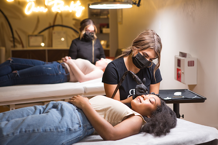 A woman with curly hair gets her eyebrows microbladed by an artist in a studio, with another person lying on a separate table in the background.