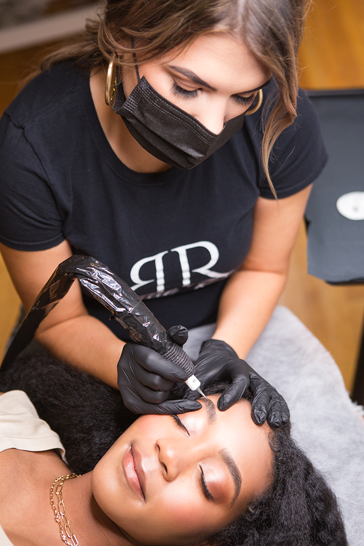 A woman with curly hair receiving a cosmetic tattoo or permanent makeup procedure on her eyebrow from a professional wearing black gloves and a face mask.