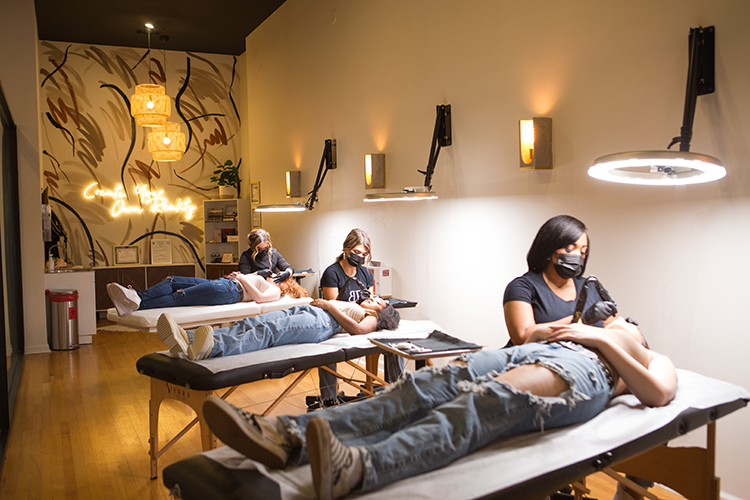 A permanent school clinic with three tables where clients are receiving microblading and other permanent cosmetic procedures, with estheticia wearing masks and working on clients. The room has modern decor, warm lighting, and a neon sign on the wall.