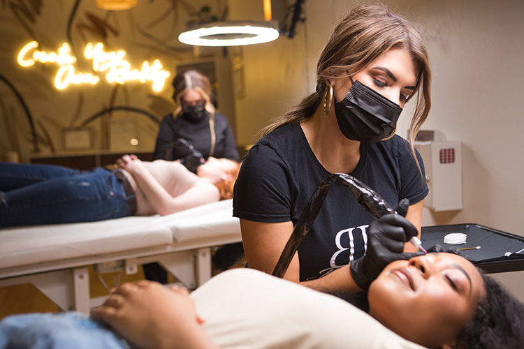 PMU artist wearing a black mask and gloves working on a client's eyebrows in a permanent cosmetics studio with another person receiving a tattoo in the background.