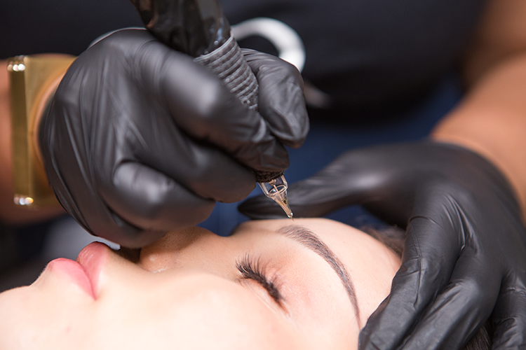 A person with closed eyes receiving a tattoo or permanent makeup procedure on their eyebrow, with technician wearing black gloves and using a tattoo machine.