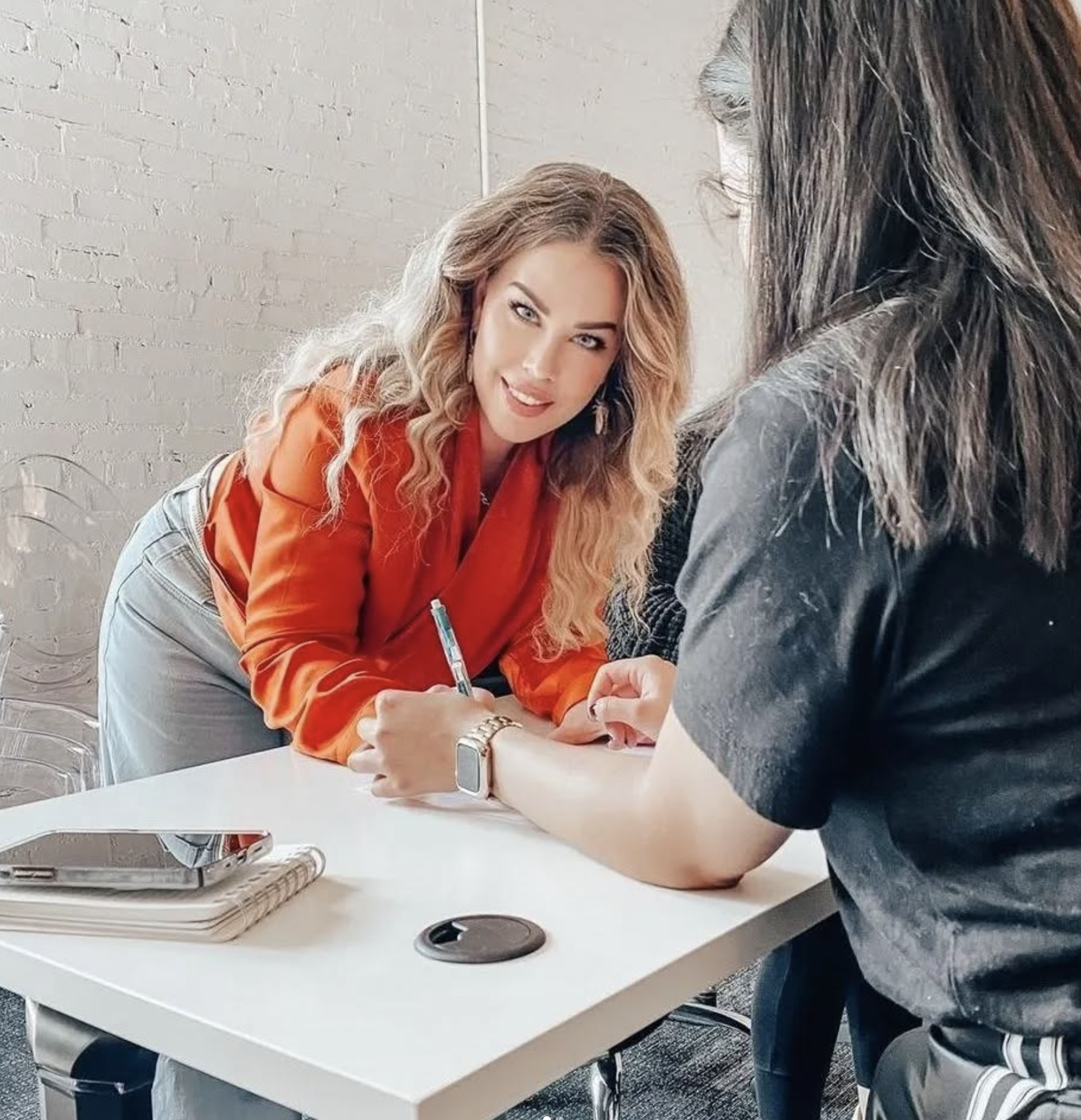 Woman in orange blazer teaching another woman in black t-shirt in a well-lit room with white brick wall.
