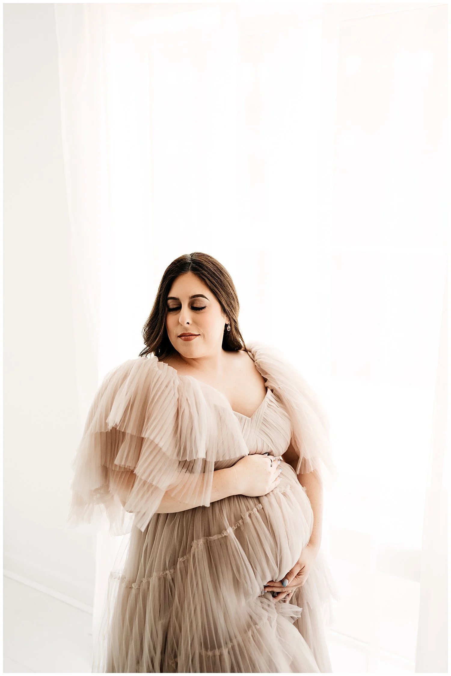 An expecting mother in a long, layered, sheer dress. Her head is turned slightly down and to the side, with her eyes shut. She cradles her belly with both hands, appearing to define it through the dress.