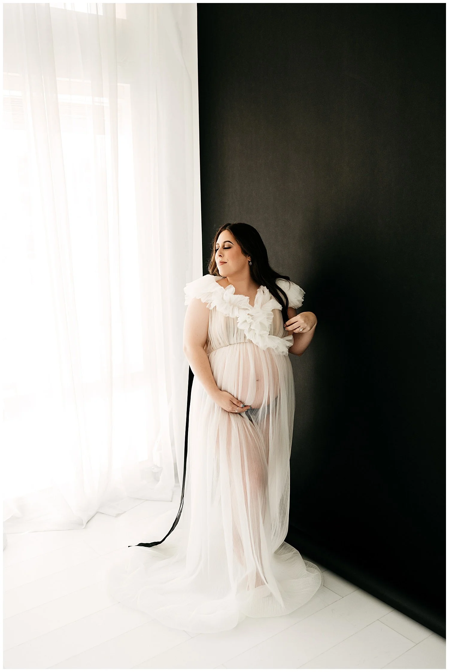 An image of an expectant mother standing in front of a window. Her dress is ruffled, sheer, and white. She turns her face upward and smiles slightly, while cradling her stomach with one hand.
