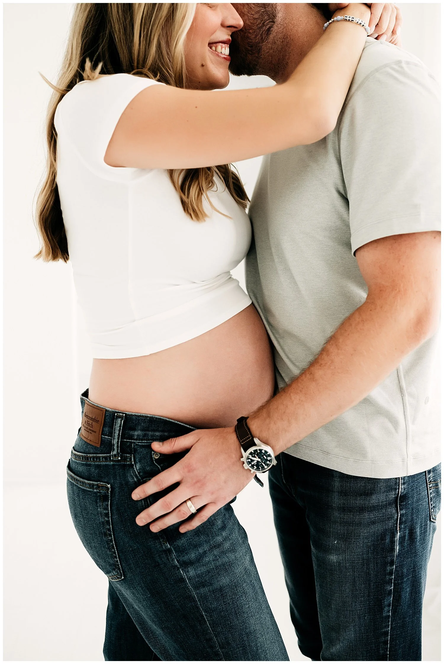 An expecting mother with her arms wrapped around her partner's neck, as he holds her pregnant belly in the embrace.