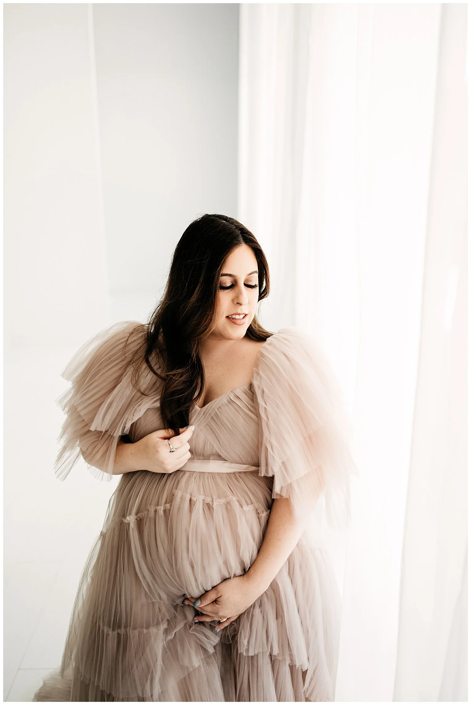 An expecting mother in a long, layered, sheer dress. Her head is turned slightly down and to the side, with her eyes shut. She cradles her belly with both hands, appearing to define it through the dress.