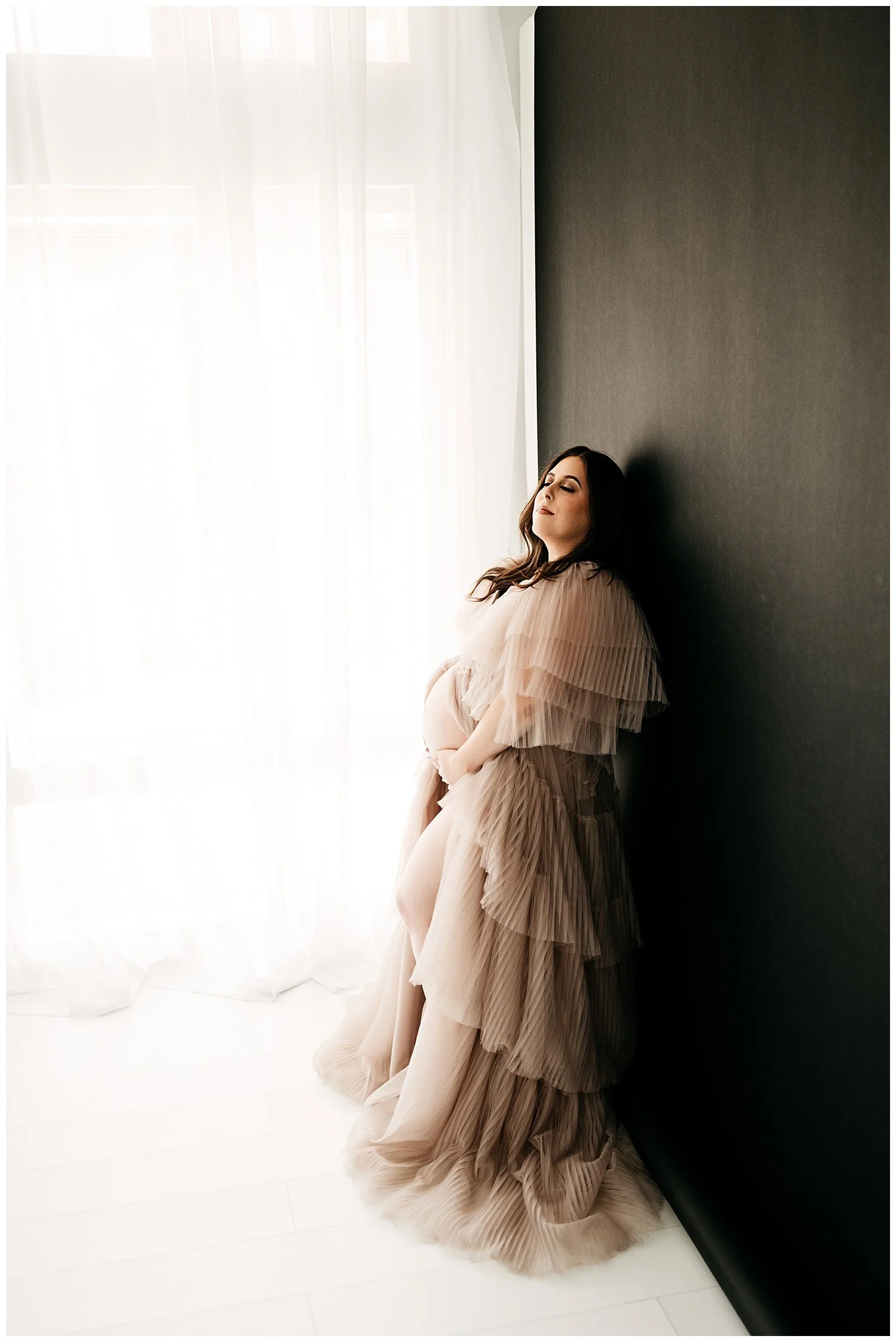 An expecting mother in a long, layered, sheer dress. Her head is turned slightly up and to the side, with her eyes shut. She cradles her belly with both hands, appearing to define it through the dress.