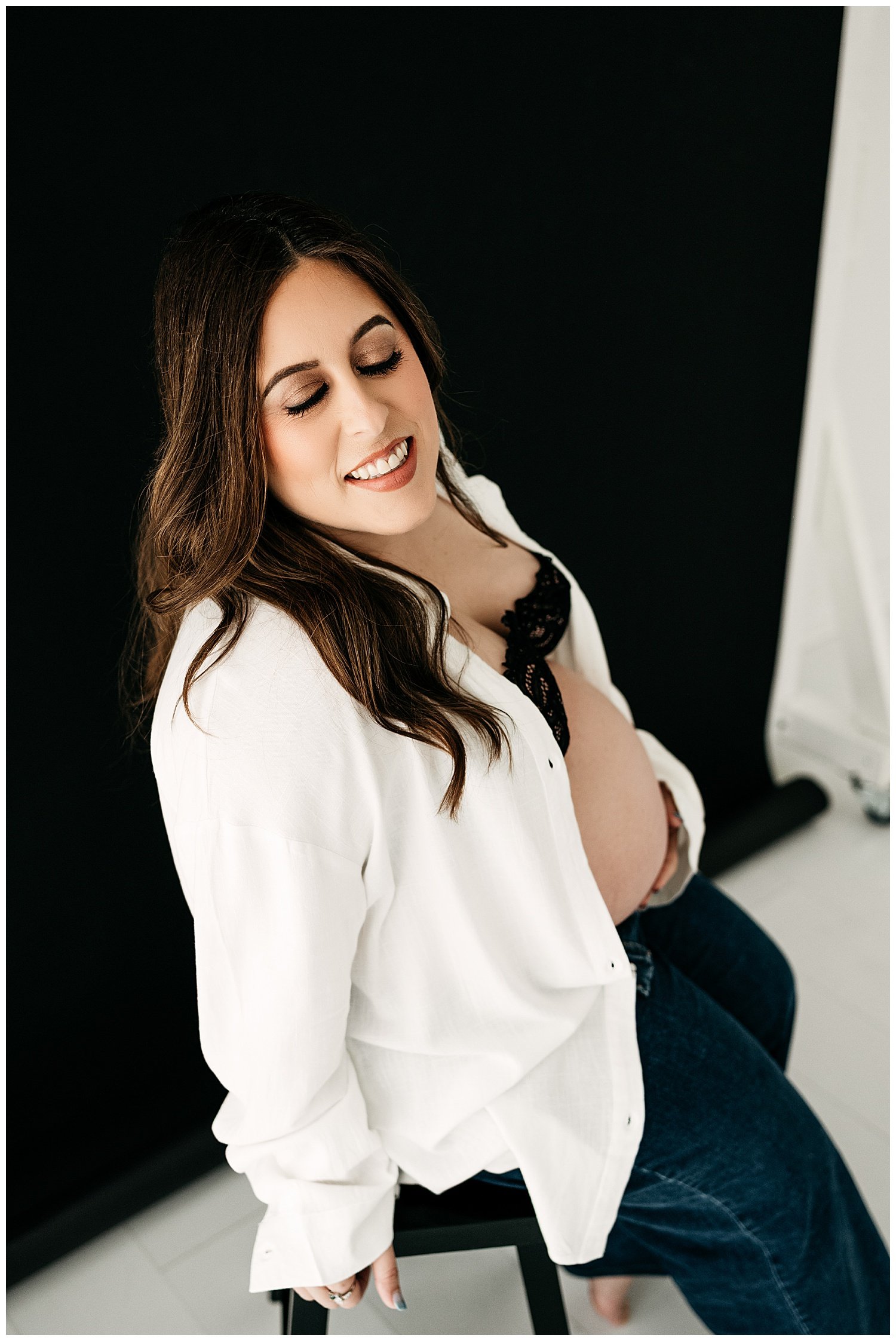 An expecting mother poses in jeans, an unbuttoned shirt, and a lacy black bra. Her stomach is exposed.