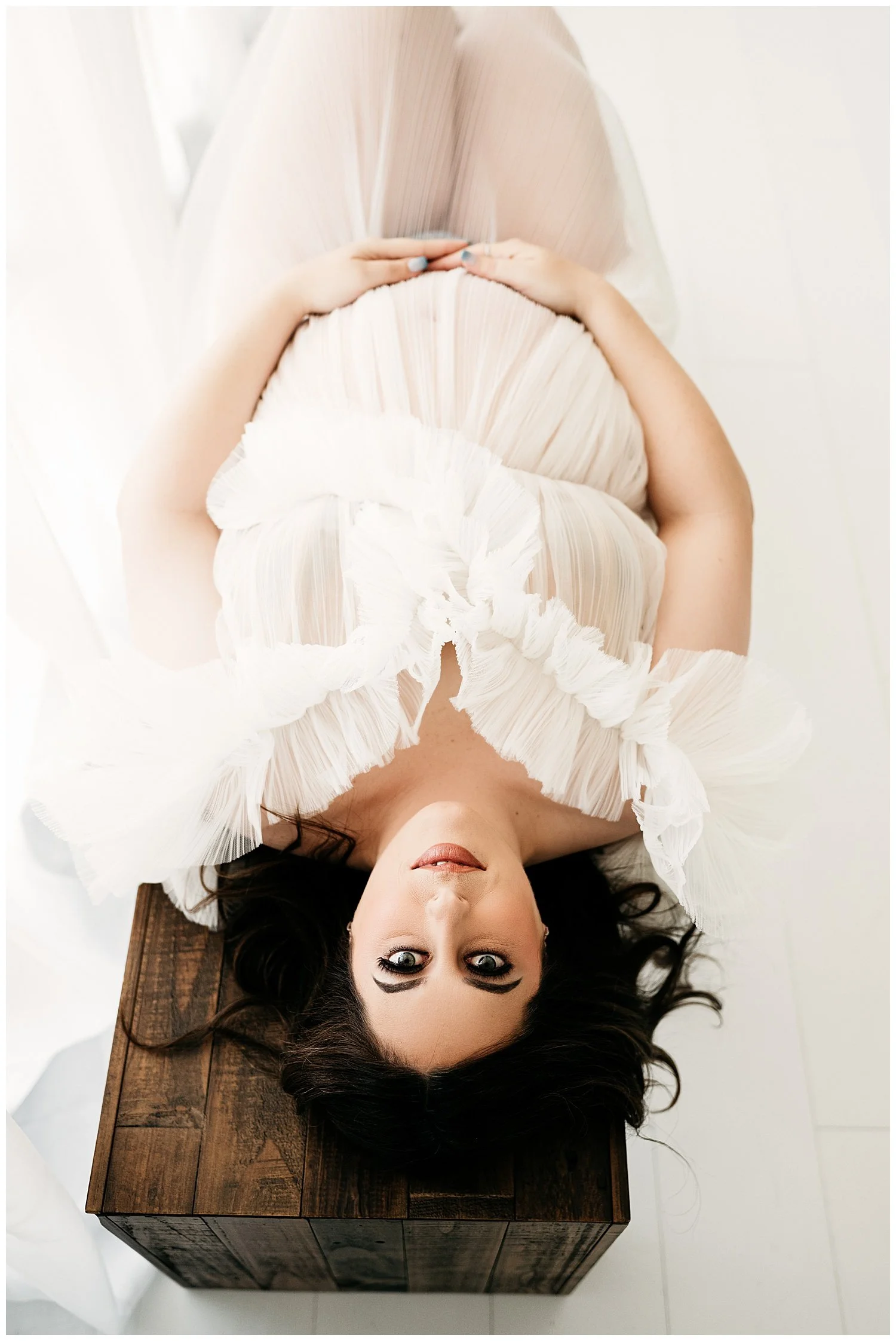 An image of an expectant mother laying a wooden bench. Her dress is ruffled, sheer, and white. She turns her face upward and smiles slightly, while cradling her stomach with one hand.