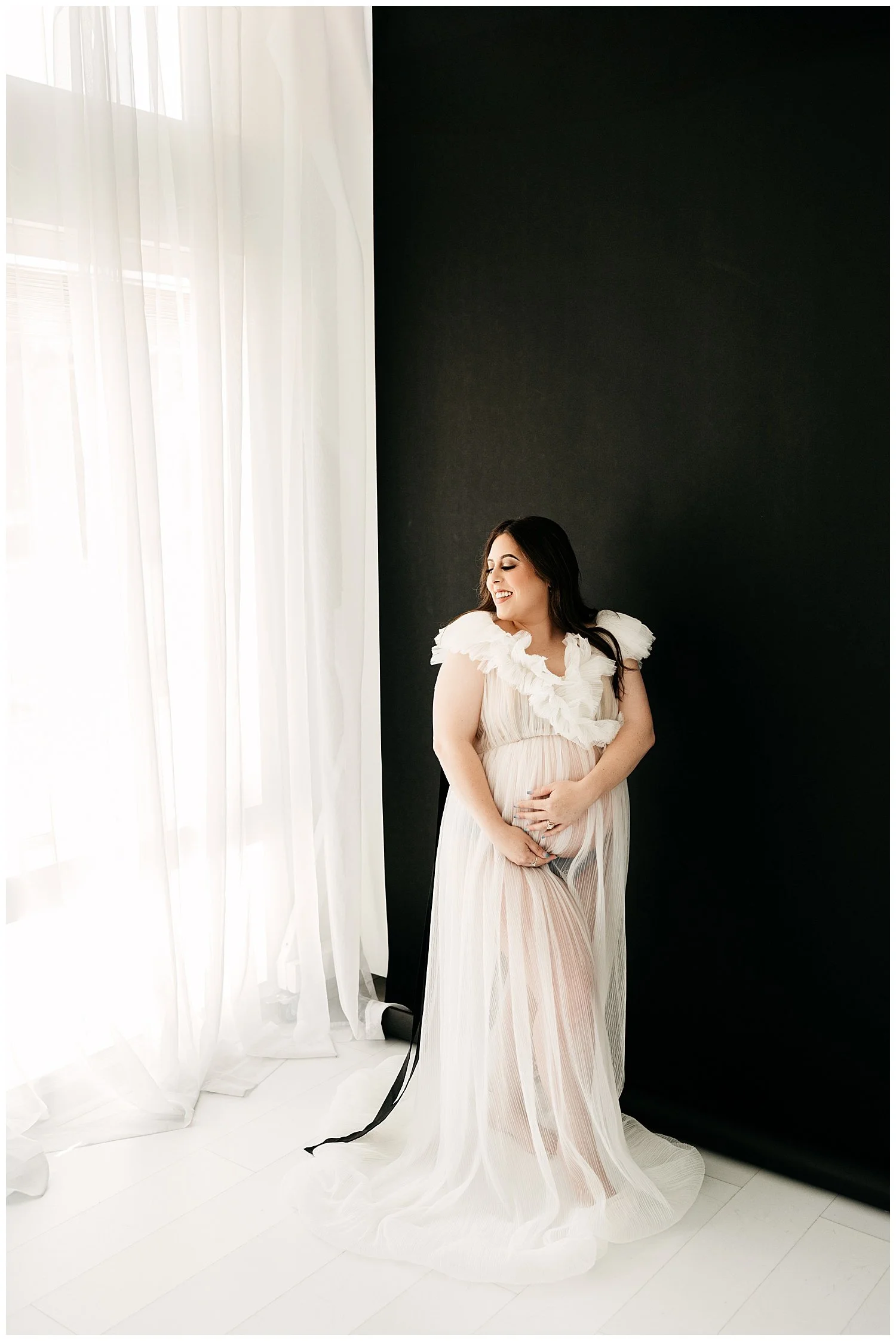 An image of an expectant mother standing in front of a window. Her dress is ruffled, sheer, and white. She turns her face upward and smiles slightly, while cradling her stomach with one hand.