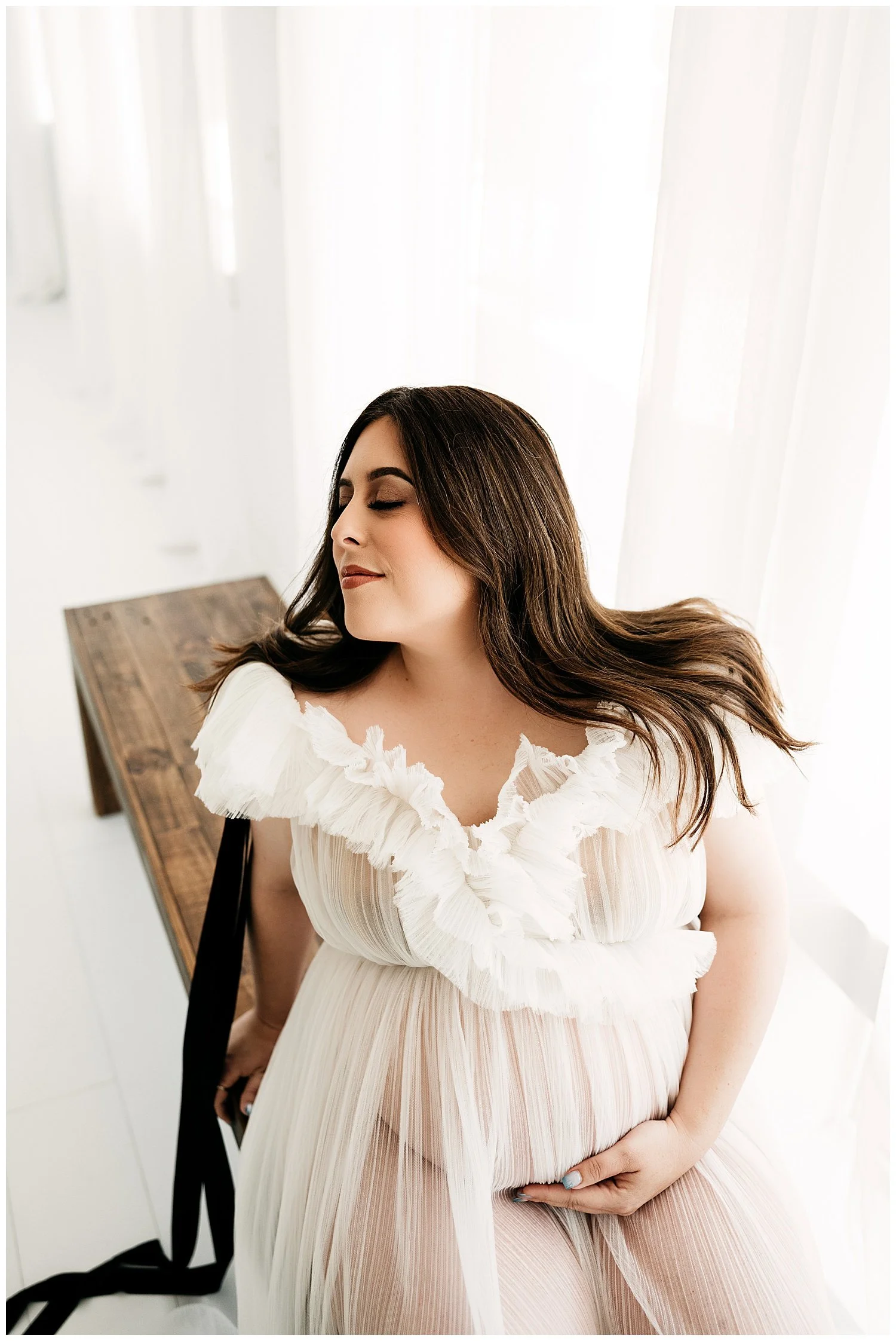 An image of an expectant mother sitting on the edge of a wooden bench. Her dress is ruffled, sheer, and white. She turns her face upward and smiles slightly, while cradling her stomach with one hand.