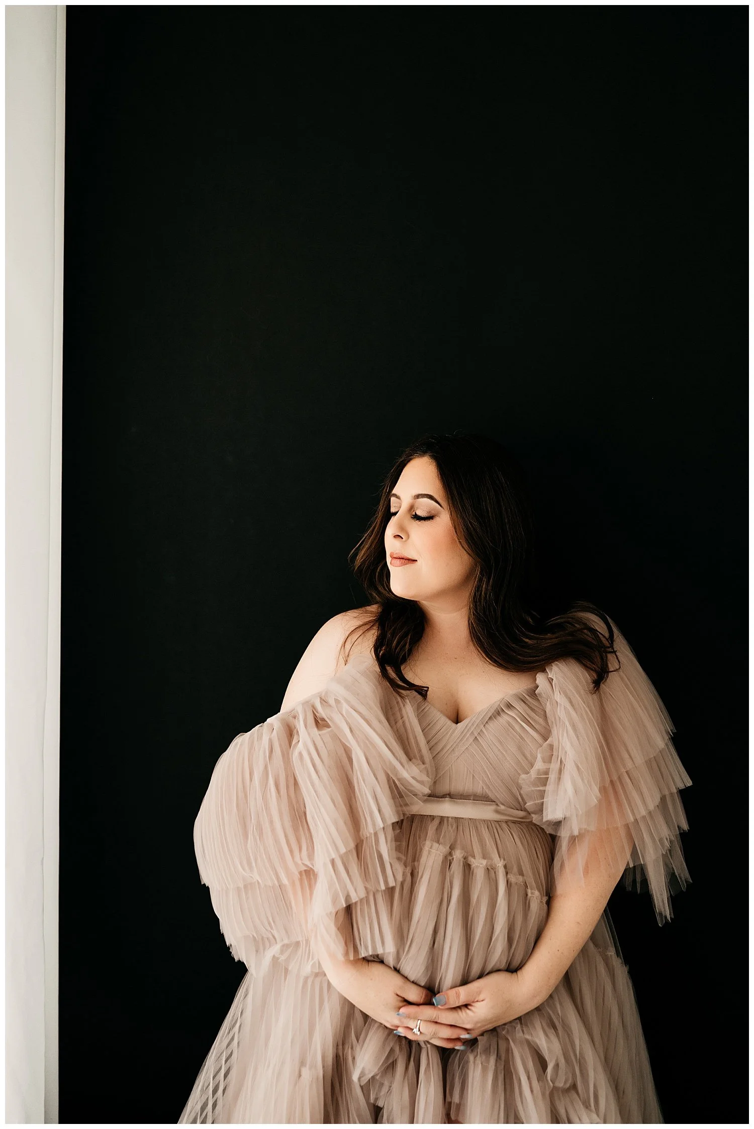 An expecting mother in a long, layered, sheer dress. Her head is turned slightly to the side, with her eyes shut. She cradles her belly with both hands, appearing to define it through the dress.