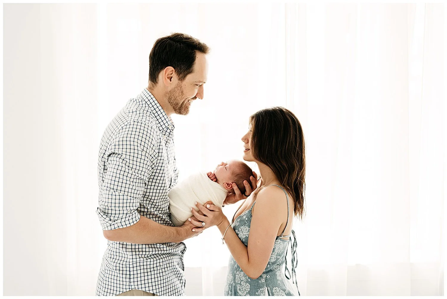An image of a couple looking lovingly into each other's eyes as they hold their newborn in between their chests.