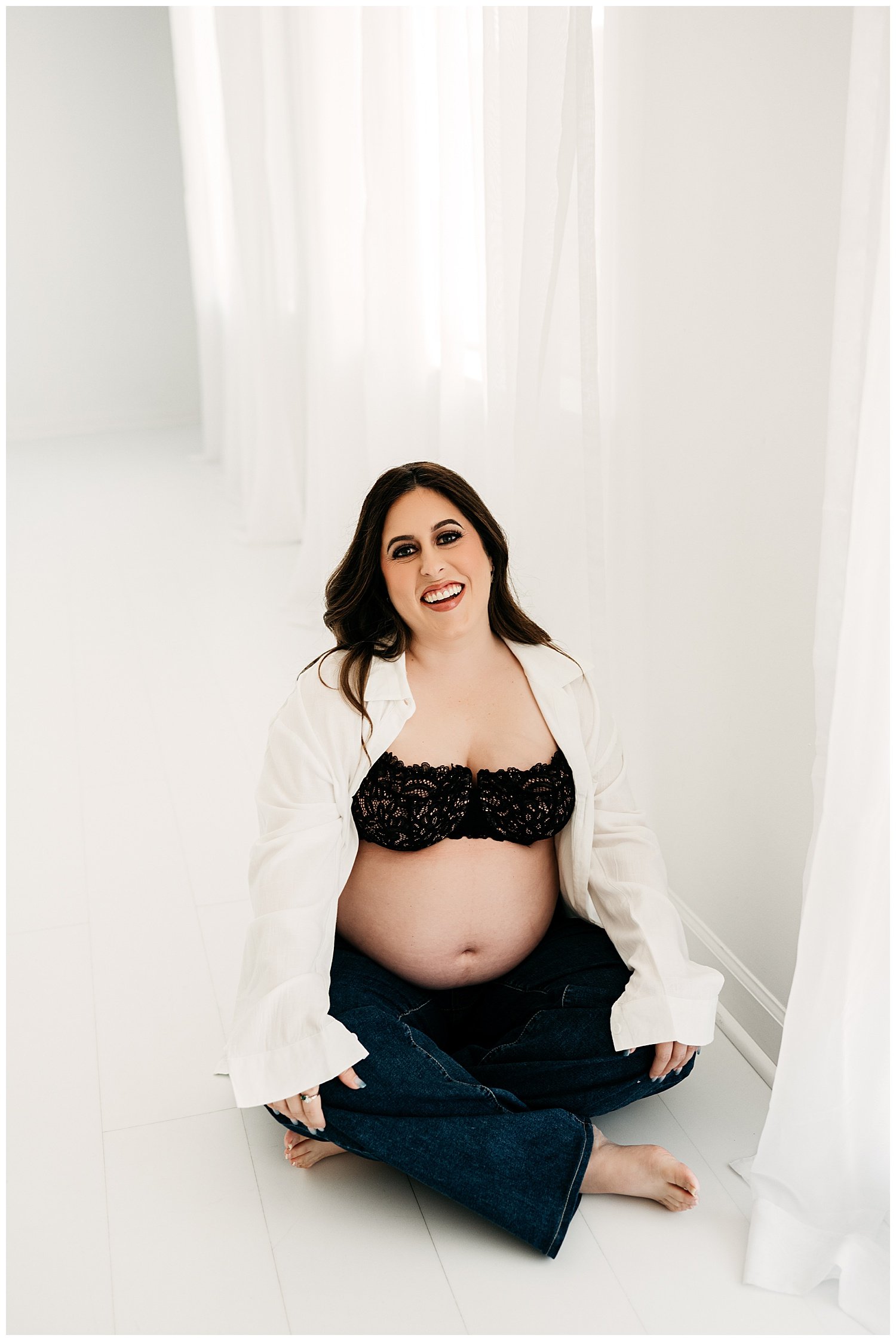 An expecting mother poses in jeans, an unbuttoned shirt, and a lacy black bra. Her stomach is exposed.