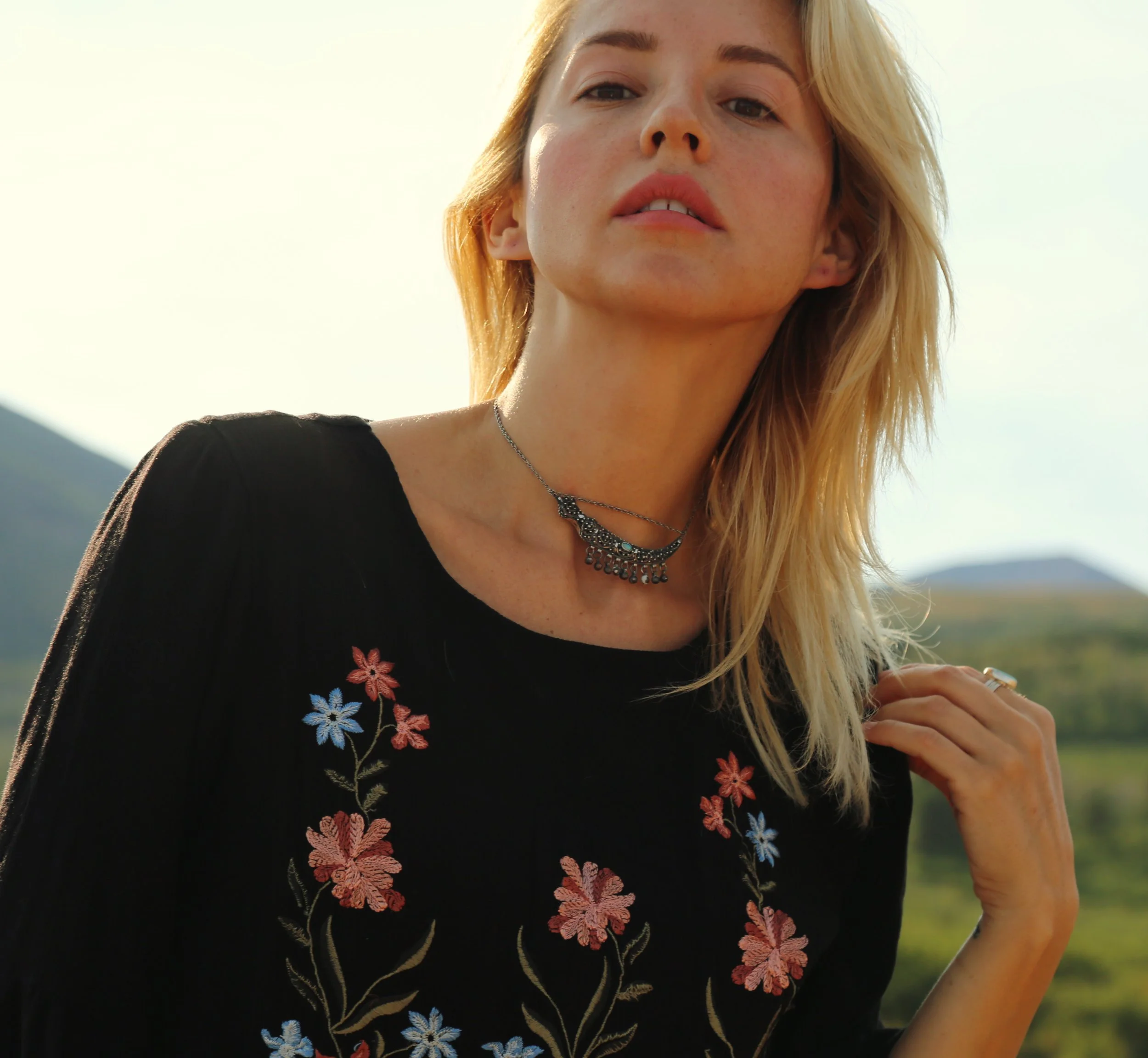 Close-up of a young woman with blonde hair, wearing a black top with colorful floral embroidery and a silver necklace, standing outdoors with a clear sky and mountains in the background.