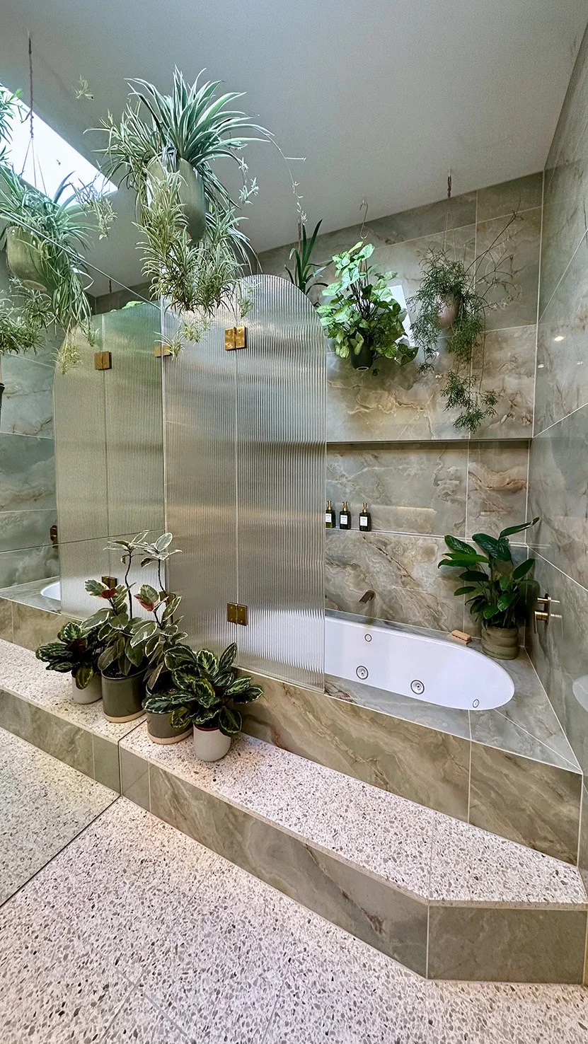 Gwendolynne Burkin interior design project. Indoor bathroom with bathtub, surrounded by beige marble tiles, potted plants, and a frosted glass privacy screen.
