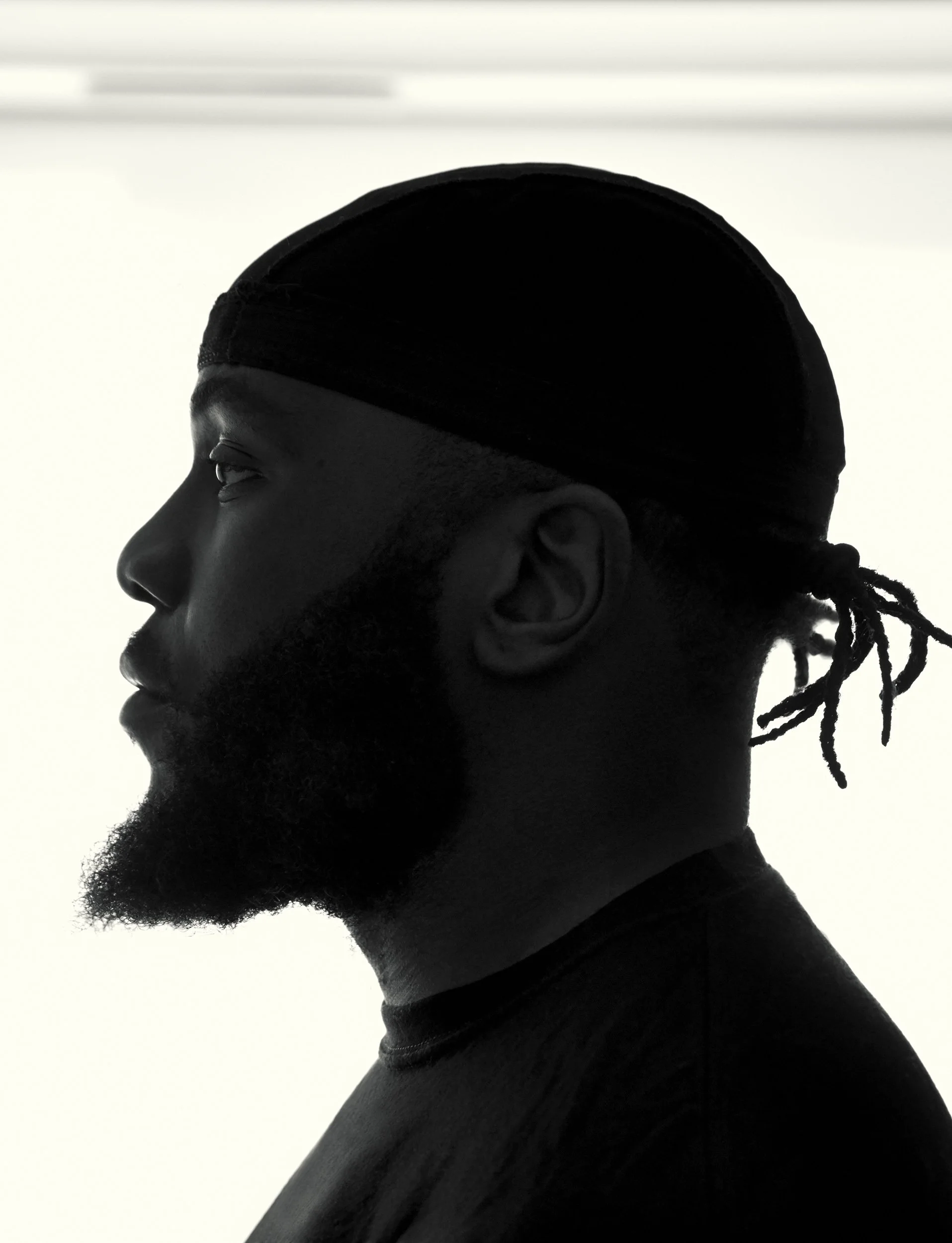 Side profile of a young man with a beard, wearing a durag, against a light background.